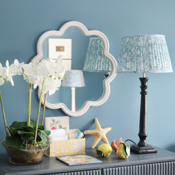 White Lacquered wavey edged shaped mirror on the wall.In front is a sideboard with an orchid plant,some stationery ceramic fruit and one of our navy lamp bases and a pleated lampshade