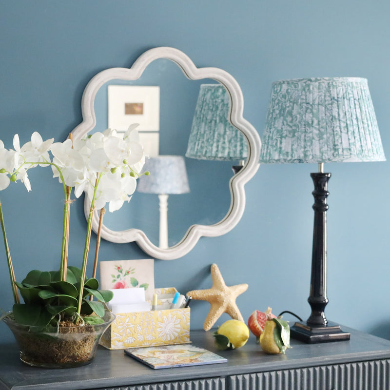 Whitewashed wooden wavey edged shaped mirror on the wall.In front is a sideboard with an orchid plant,some stationery, ceramic fruit and one of our navy lamp bases and a pleated lampshade