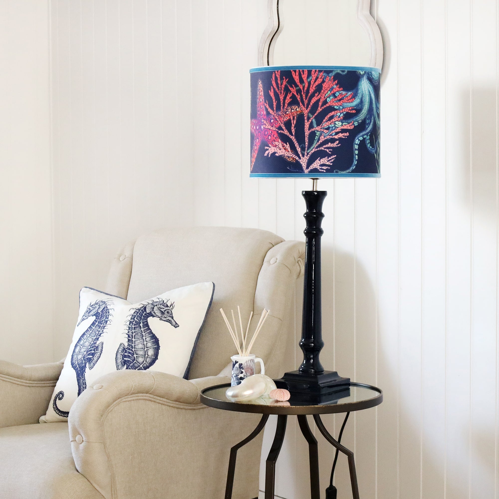 Navy Lacquer Godolphin lampbase  with a navy reef lampshade on a mirror table.On the table is a beachcomber diffuser with some shells placed on it.Next to the table is a chair with a seahorse cushion