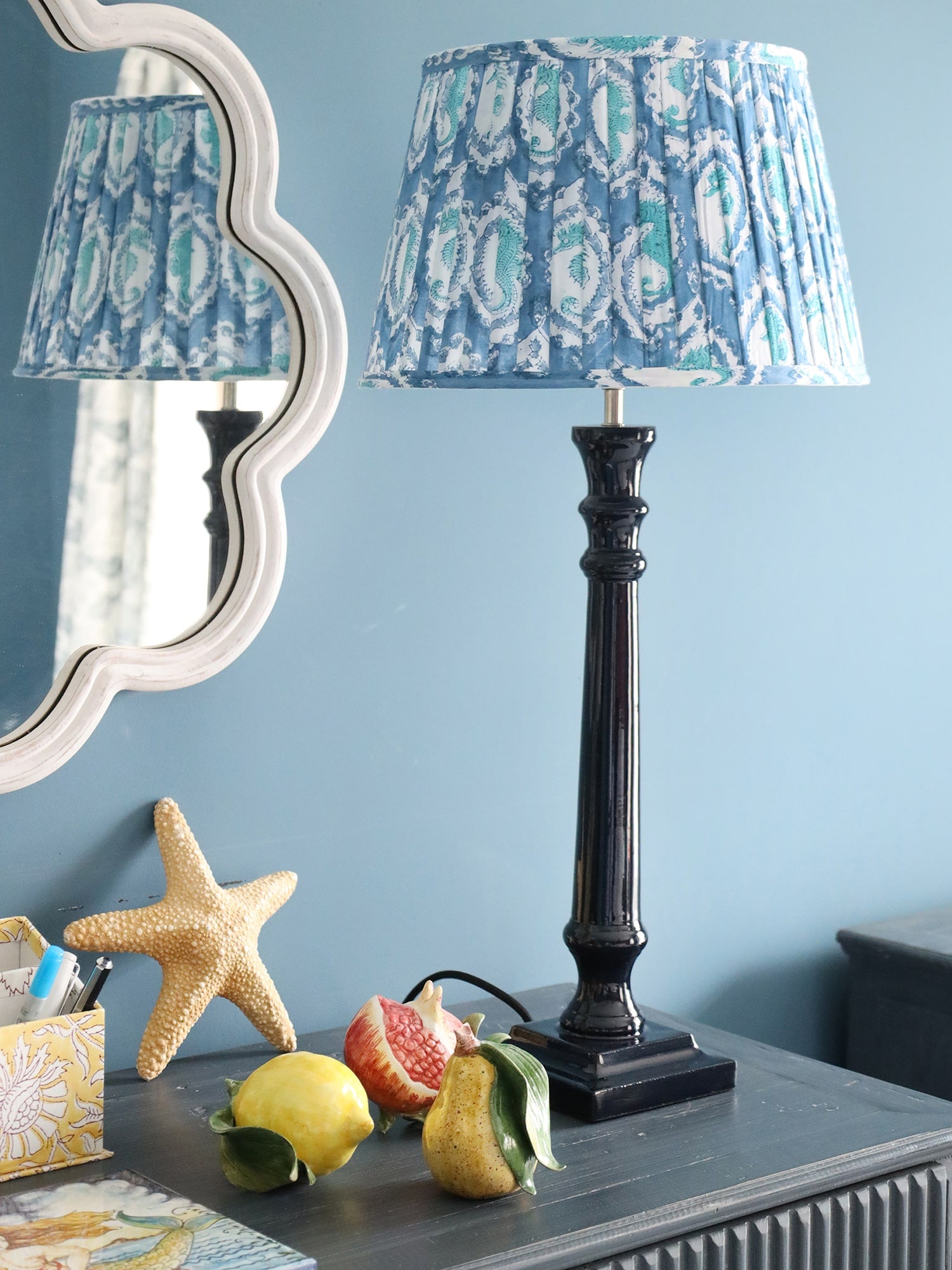 Large Azure paisley pleated lampshade on our Navy lampbase placed on a sideboard.On the sideboard are ceramic fruits and a starfish,in the background is a white wave mirror.