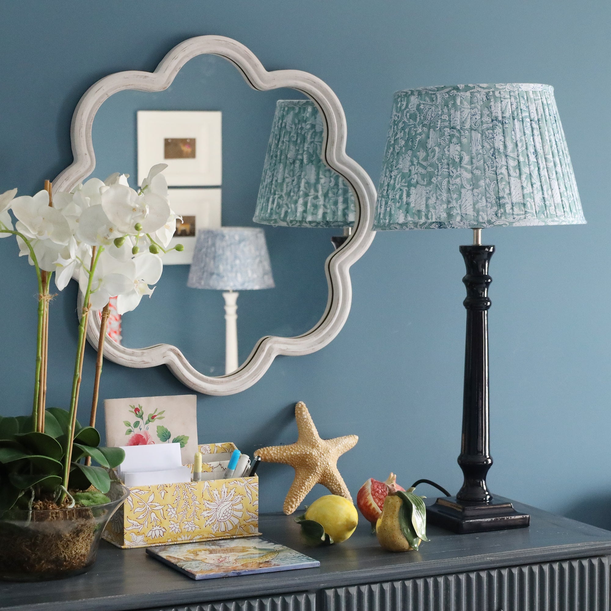 Navy Lacquer Godophin lampbase with a pleated lampshade on a sideboard.On the sideboard are several ceramic fruits,staionary, an orchid in a pot and a starfish.Behind on the wall is a wave mirror 