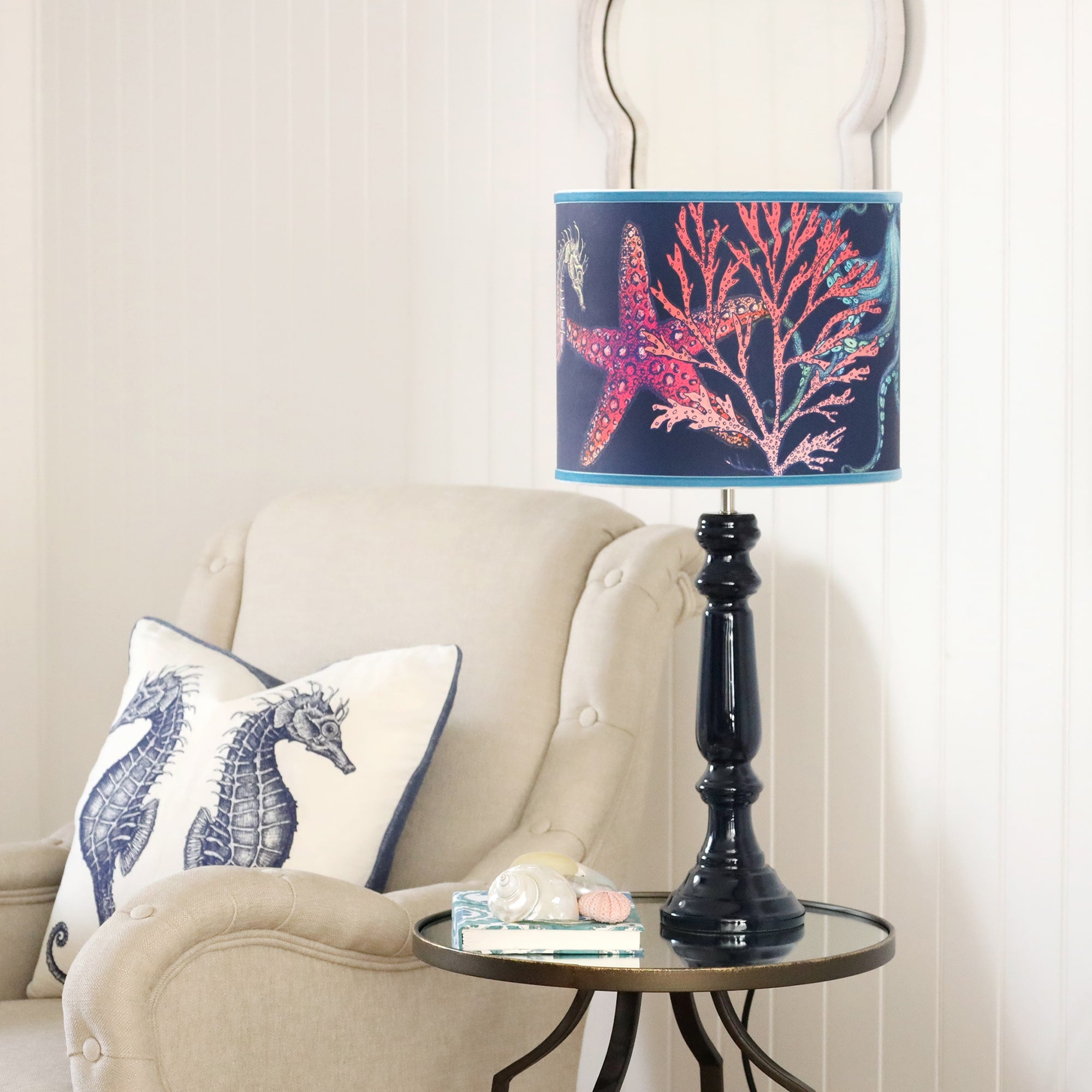 Navy Lacquer Zennor lampbase with a navy reef lampshade on a mirror table.On the table is a notebook with some shells placed on it.Next to the table is a chair with a seahorse cushion
