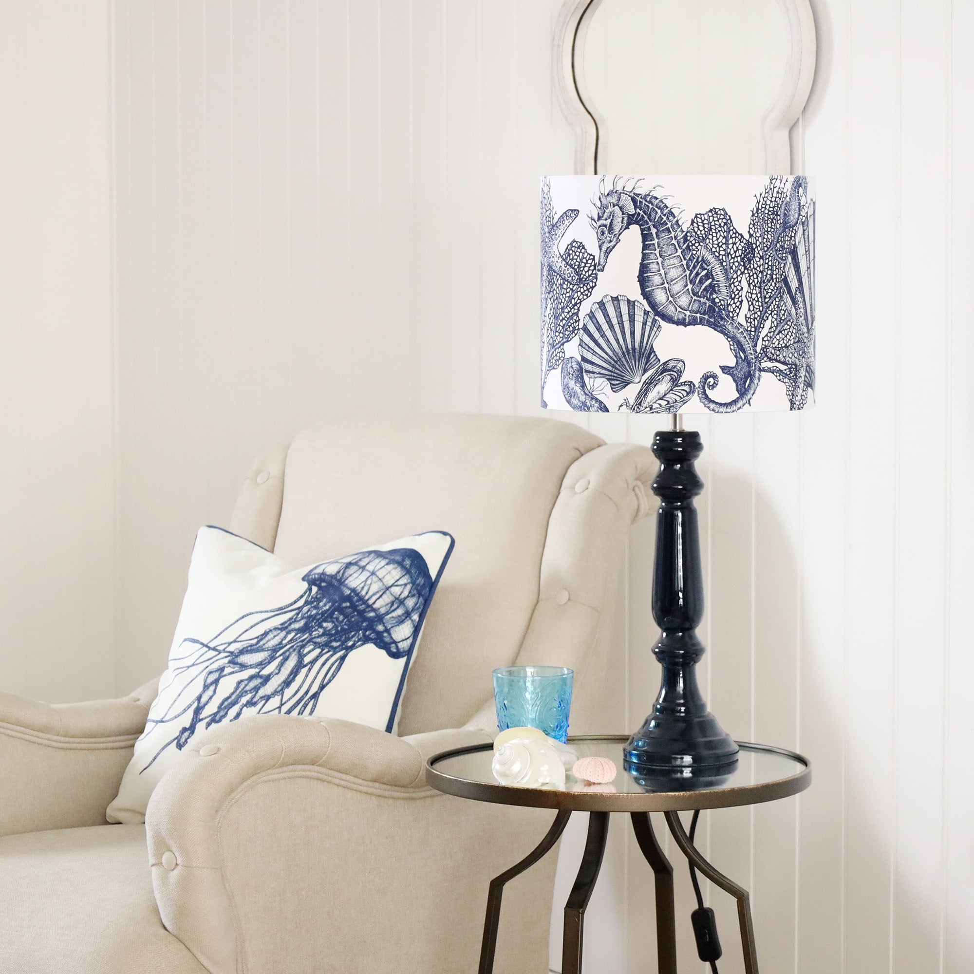 Navy Lacquer Zennor lamp base with a seahorse lampshade on a mirror table.On the table is a glass with some shells placed on it.Next to the table is a chair with an jellyfish cushion.Behind on the wall is a wave mirror.