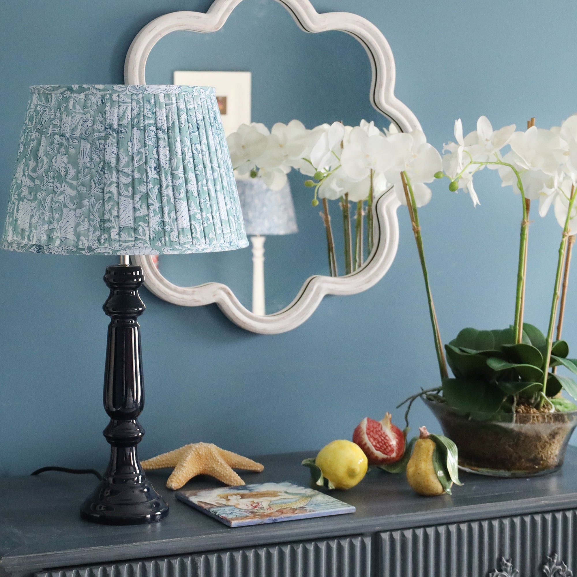 Navy Lacquer Zennor lampbase with a pleated lampshade on a sideboard.On the sideboard are several ceramic fruits,stationery, an orchid in a pot and a starfish.Behind on the wall is a wave mirror