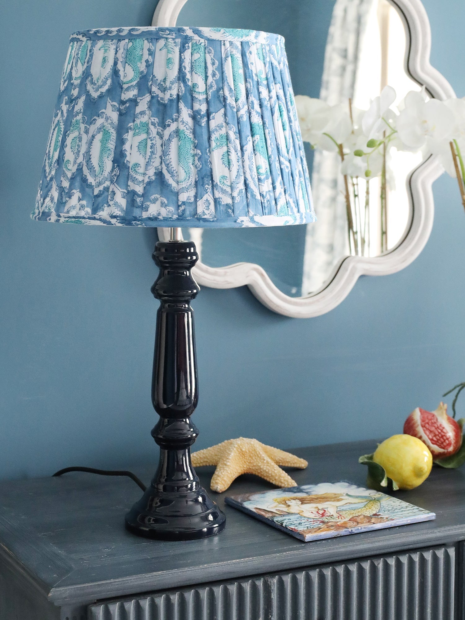 Navy Lacquer Zennor lampbase with a pleated lampshade on a sideboard.There are ceramic fruits and a starfish,on the wall behind is a wave mirror.