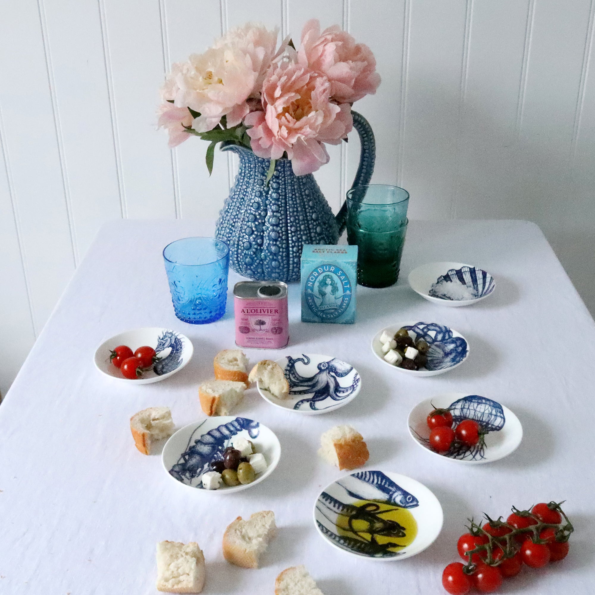 Nibbles bowl in Bone China in our Classic range in Navy and white in the Seahorse design placed on a white table cloth are more nibbles bowls with various foodstuffs in,in the background is a large blue jug with flowers and colourful glasses