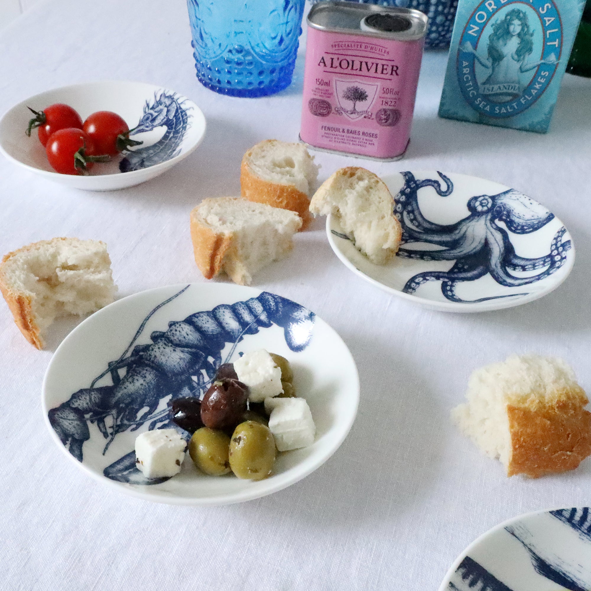 Nibbles bowl in Bone China in our Classic range in Navy and white in the Lobster design placed on a white table cloth are more nibbles bowls with various foodstuffs in,in the background is a large blue jug and colourful glasses