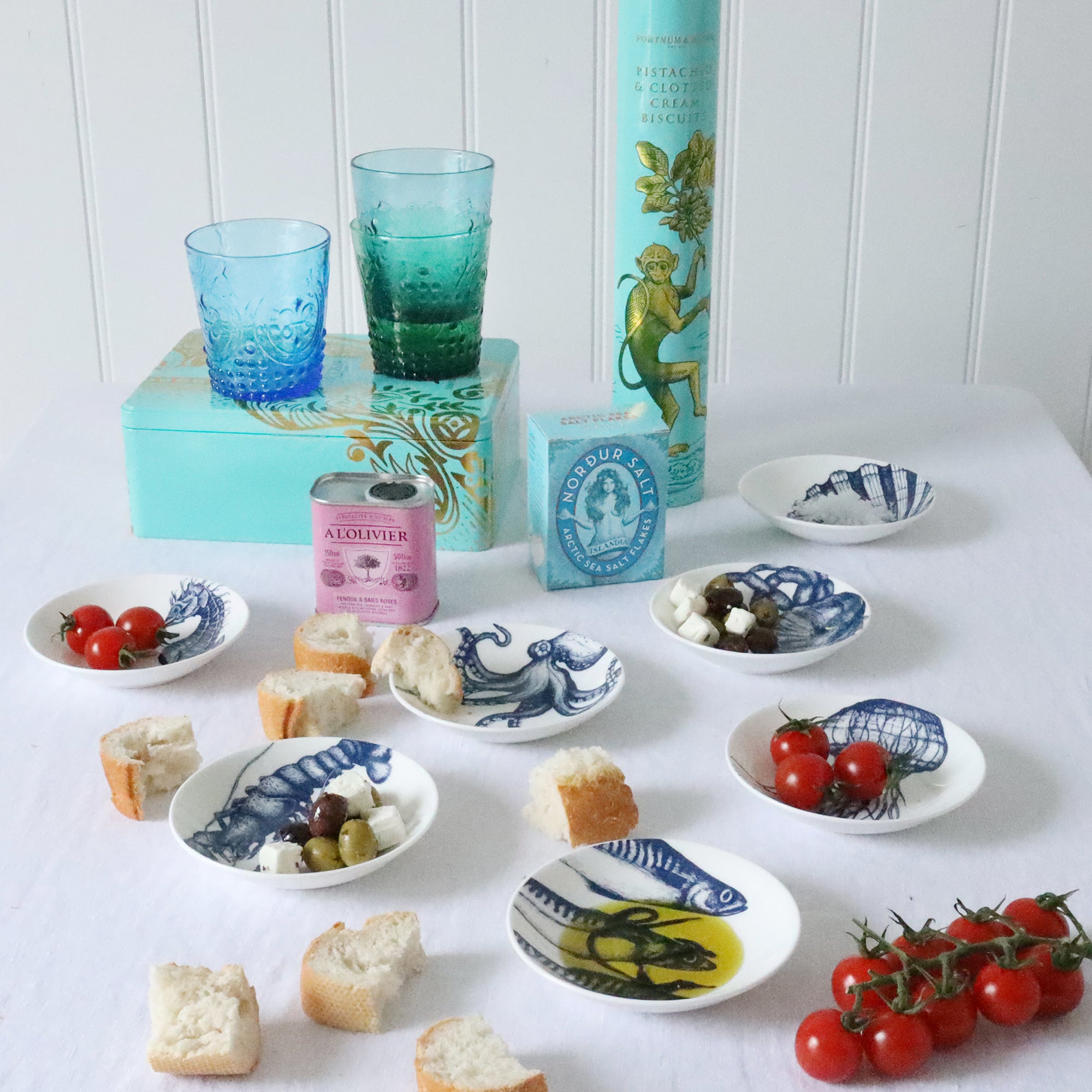 Nibbles bowl in Bone China in our Classic range in Navy and white in the Scallop design placed on a white table cloth are more nibbles bowls with various foodstuffs in,in the background are a couple of  large blue biscuit tins and colourful glasses