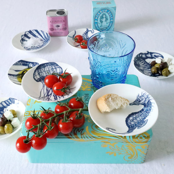 Nibbles bowl in Bone China in our Classic range in Navy and white in the Seahorse design placed on a turquoise box with cherry tomatoes.Also on the box is a Mackerel nibbles dish with some bread in it.On the table below on a white table cloth are more nibbles bowls with various foodstuffs in