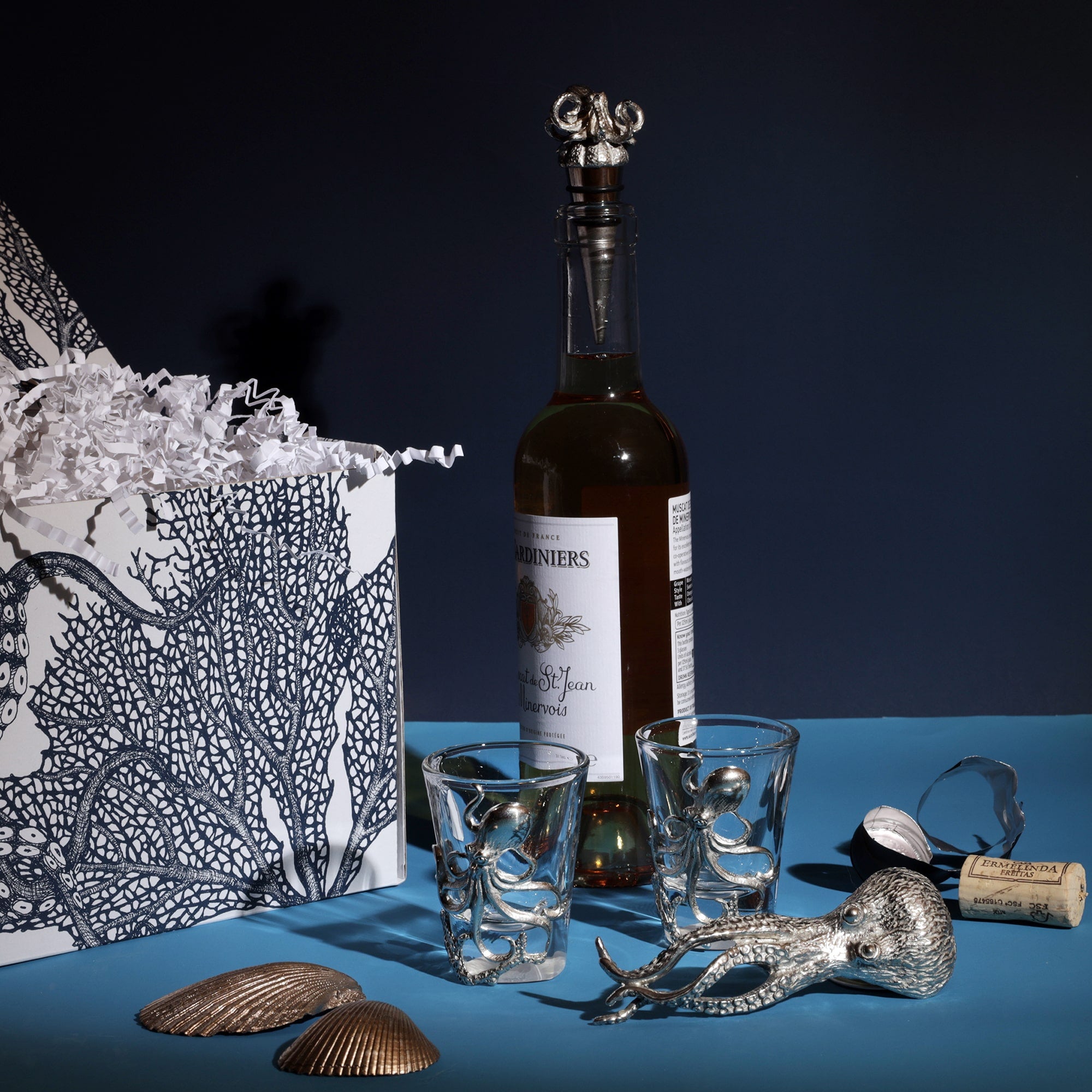 A Cream Cornwall gift set featuring an octopus-themed wine stopper, two shot glasses with octopus designs, and a bottle of wine. The items are displayed next to a decorative gift box adorned with octopus and coral illustrations. Decorative seashells are scattered around the set, enhancing the coastal theme against a dark blue background.