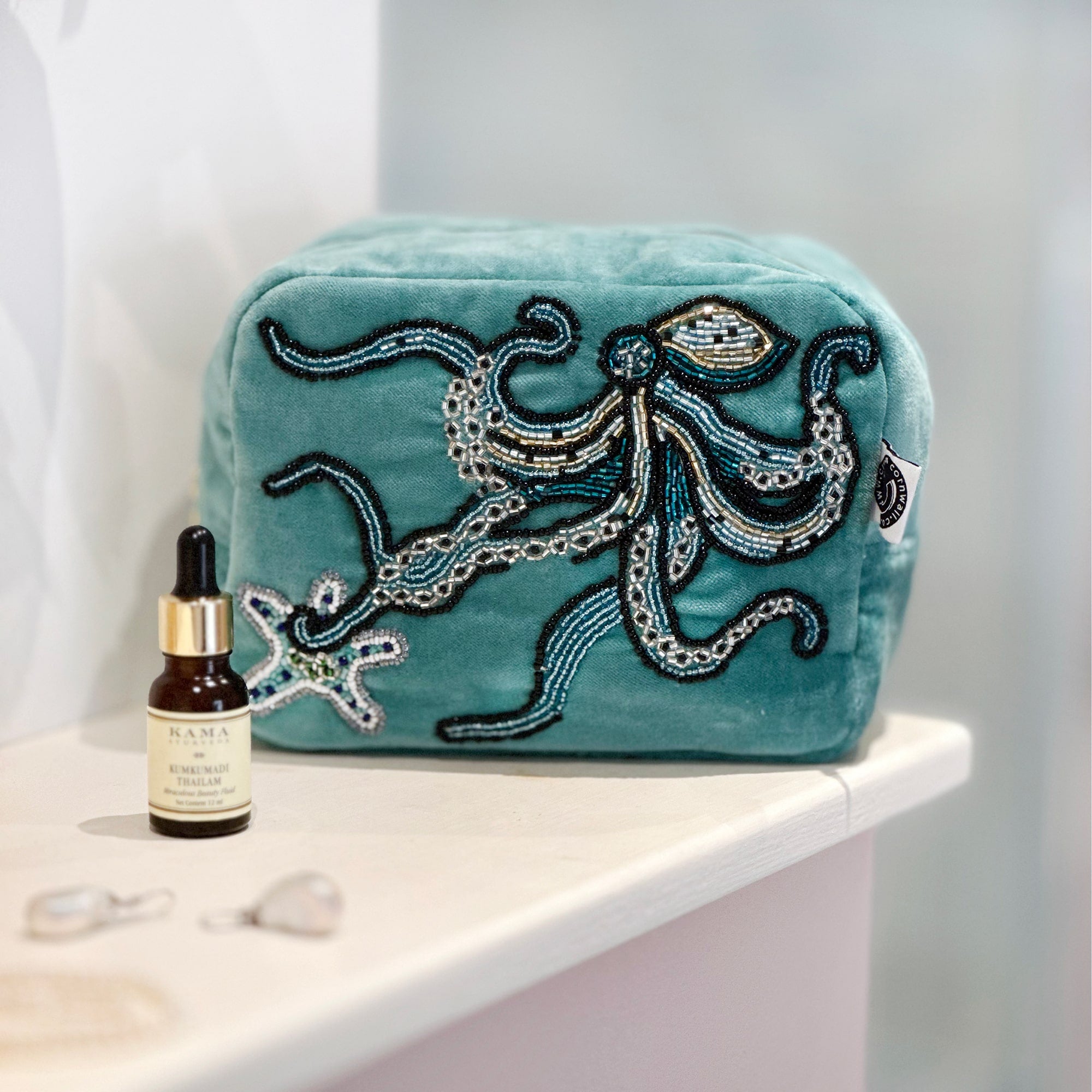 Beaded aqua velvet cosmetic bag with octopus and starfish decoration sitting on a bathroom shelf with a small glass bottle pipette and some pearl earrings in front.