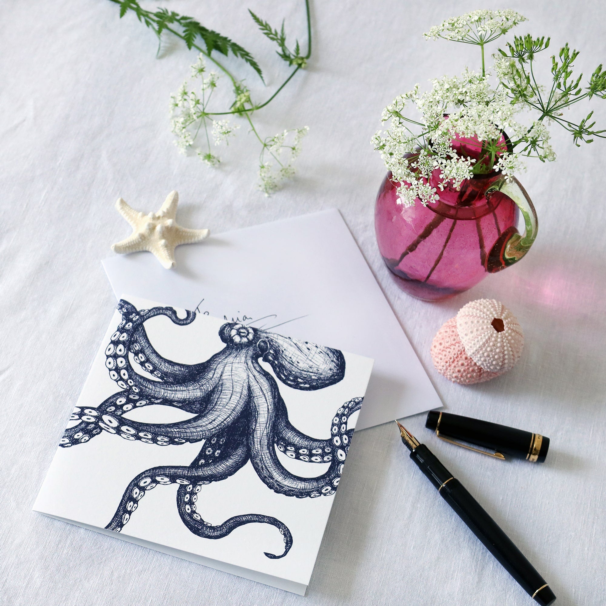 greeting card with navy blue illustrated octopus that looks like it is dancing across the card lying on a white table cloth with a fountain pen, hand written envelope shells and a small cranberry glass jug with wild flowers in