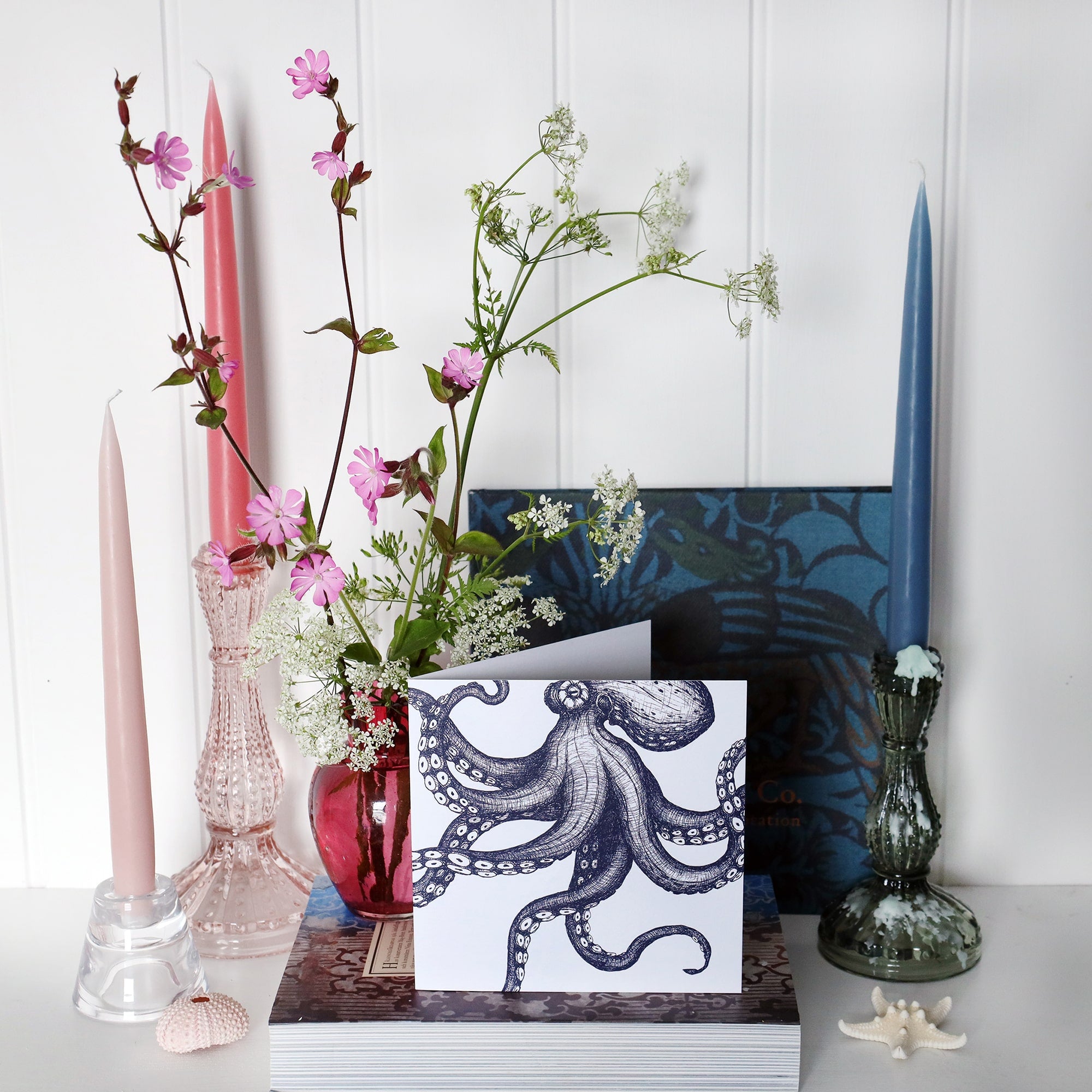 greeting card with navy blue illustrated octopus that looks like it is dancing across the card on shelf with pink and blue candles in candlesticks and a small cranberry glass jug with wild flowers in