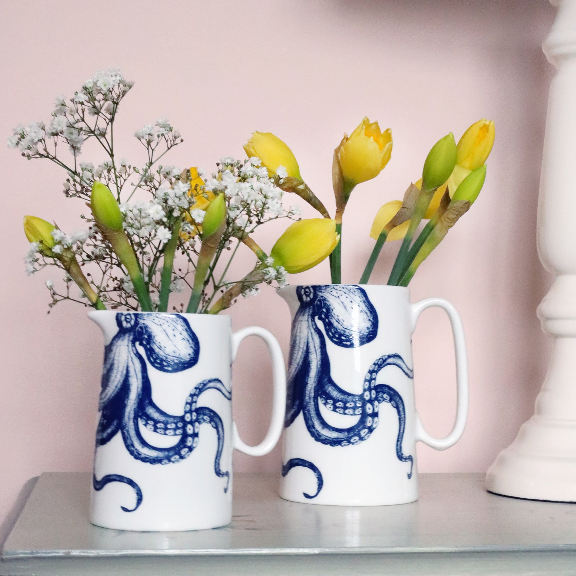 2 white jugs with a navy illustration of an octopus on them. they have yellow daffodils in them and gypsophila with a soft pink wall behind them and the edge of white lamp base just visible.