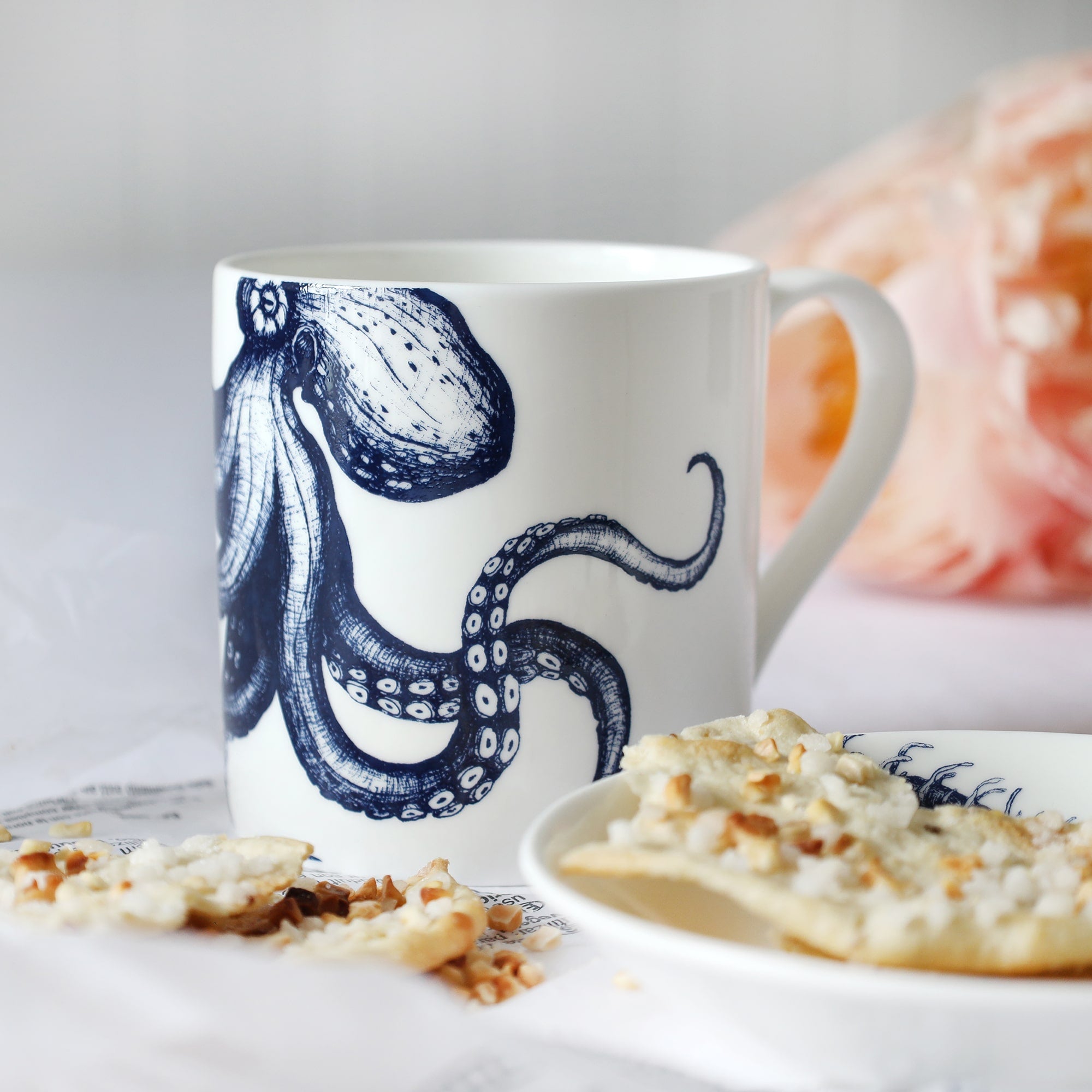 White mug with dark blue octopus illustration sitting on a table with a plate & biscuits and flowers in the background.