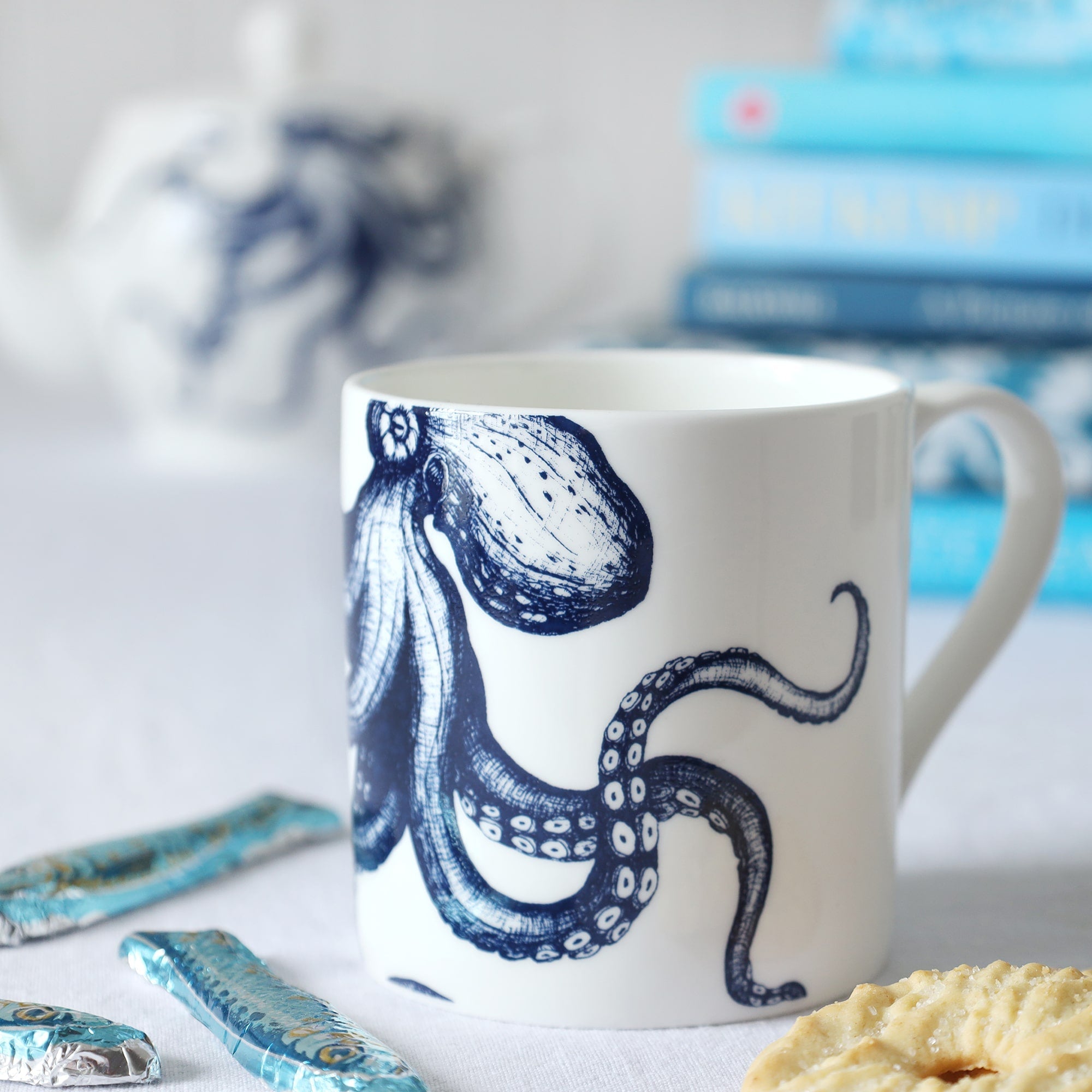 Close up of a white bone china mug with a blue illustrated octopus on it, sitting on a table with biscuits and sardine chocolates and a pile of out of focus blue books and teapot in the background. 
