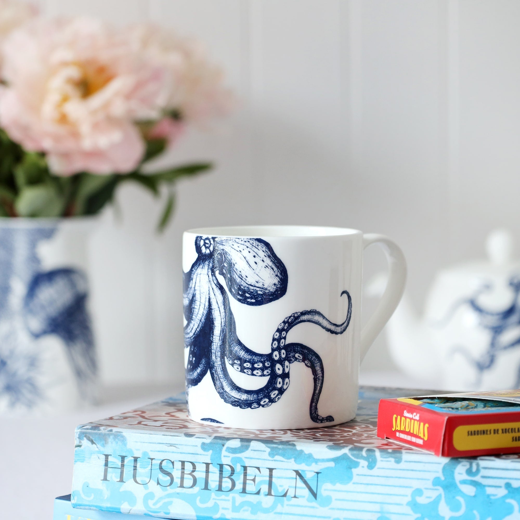 Close up of a white bone china mug with a blue illustrated octopus on it, sitting on a book with chocolate sardines and out of focus teapot & flowers in the background. 