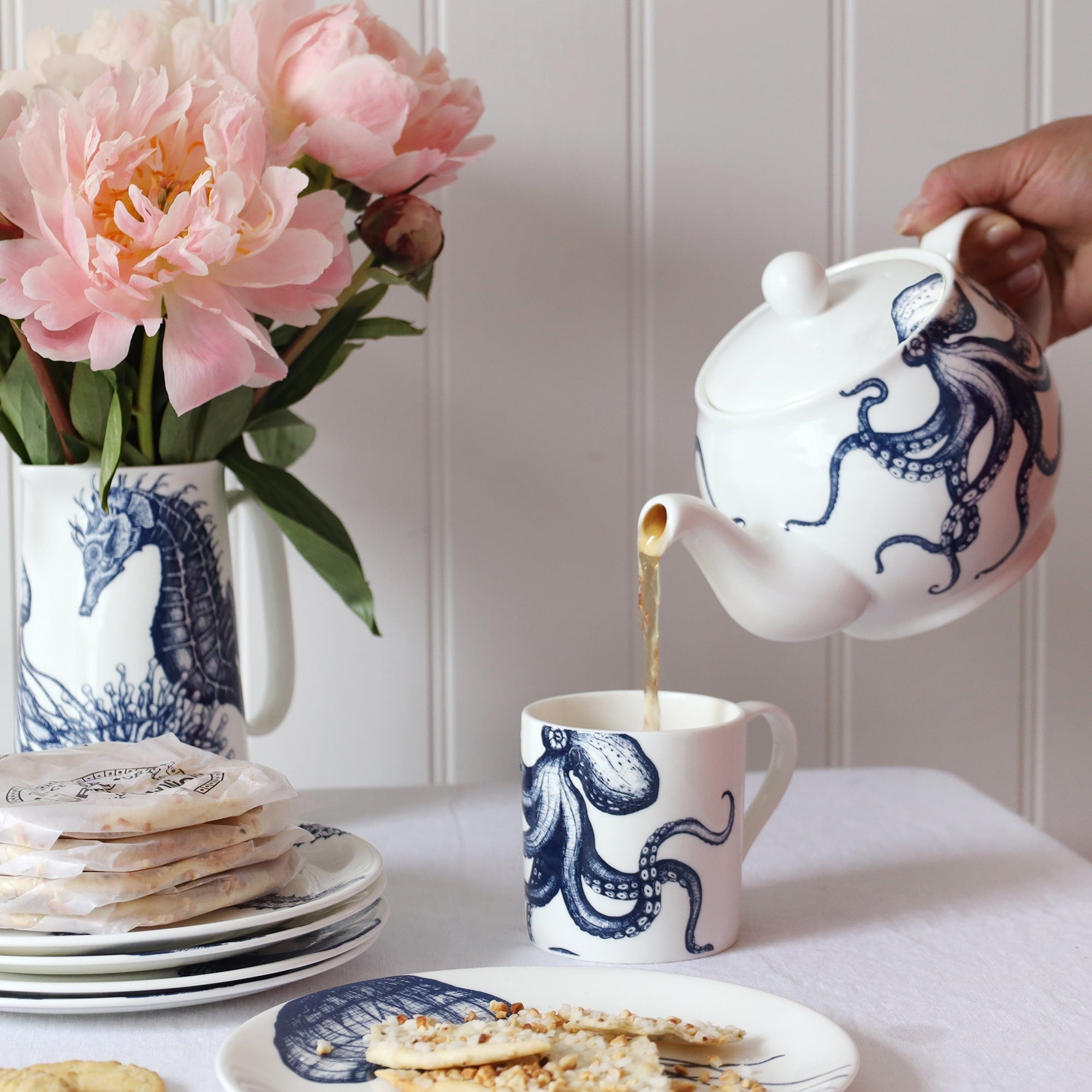 A blue & white informal table setting set against a white shiplap wall and linen tablecloth. An octopus design adorns a teapot which is pouring tea into a white bone china mug decorated with a blue illustrated octopus..