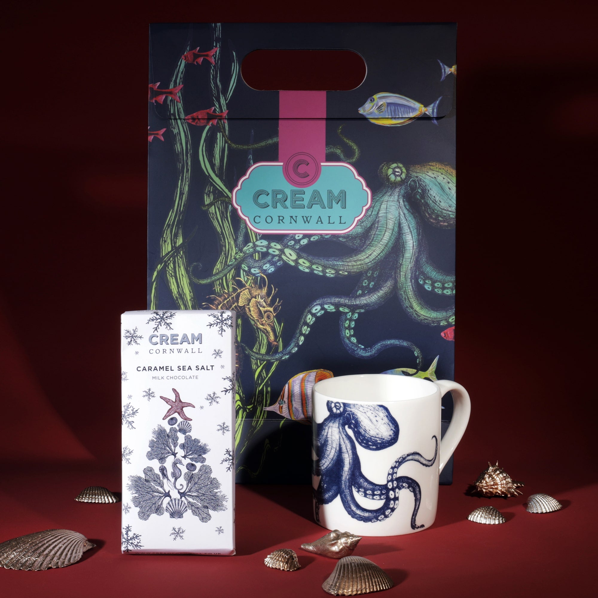 A Cream Cornwall gift set featuring a ceramic mug with a blue octopus design and a bar of caramel sea salt chocolate. The items are displayed alongside a colorful marine-themed gift bag decorated with illustrations of an octopus, fish, and seaweed. Decorative seashells are scattered around the products, enhancing the coastal theme against a deep red background.