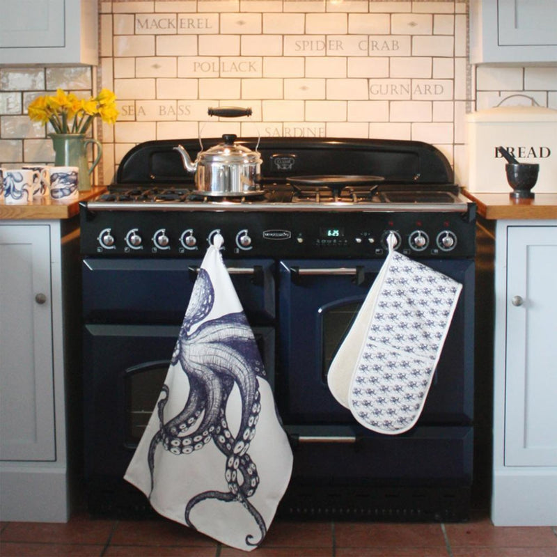 Cotton and Linen Octopus Tea towel decorated with hand drawn design in shades of blue on a white background hanging from an aga handle.On the aga is a shiny silver kettle,also hanging is one of our oven gloves and on the side are several of our mugs in front of a jug full of daffodils.On the other side is a pestle and mortar in front of a bread bin