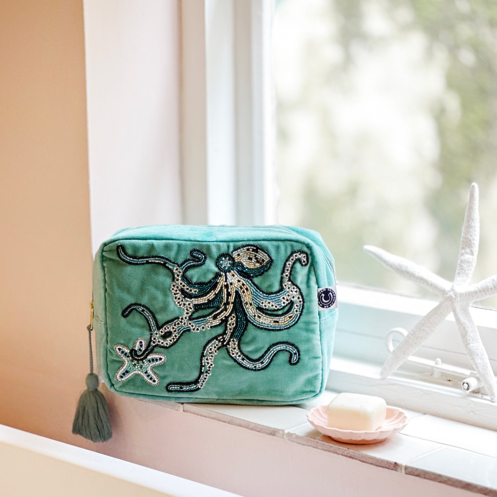 Beaded aqua velvet wash bag with octopus and starfish decoration sitting in a bathroom window with the sun coming in and a shell soap dish and starfish next to it.