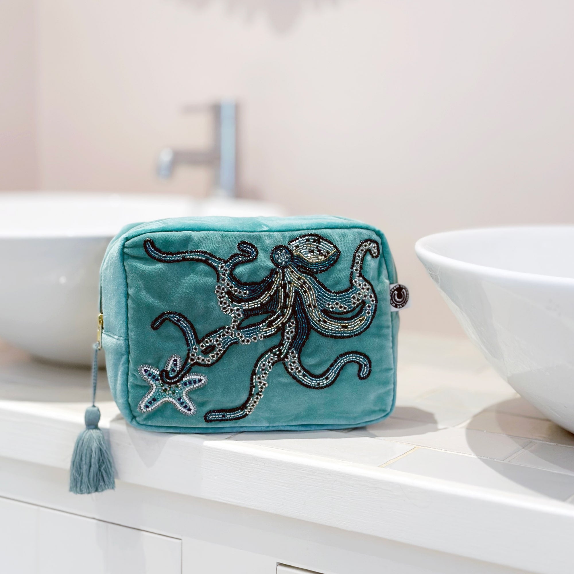 Beaded aqua velvet wash bag with octopus and starfish decoration on a tiled bathroom top in between 2 round white wash basins.