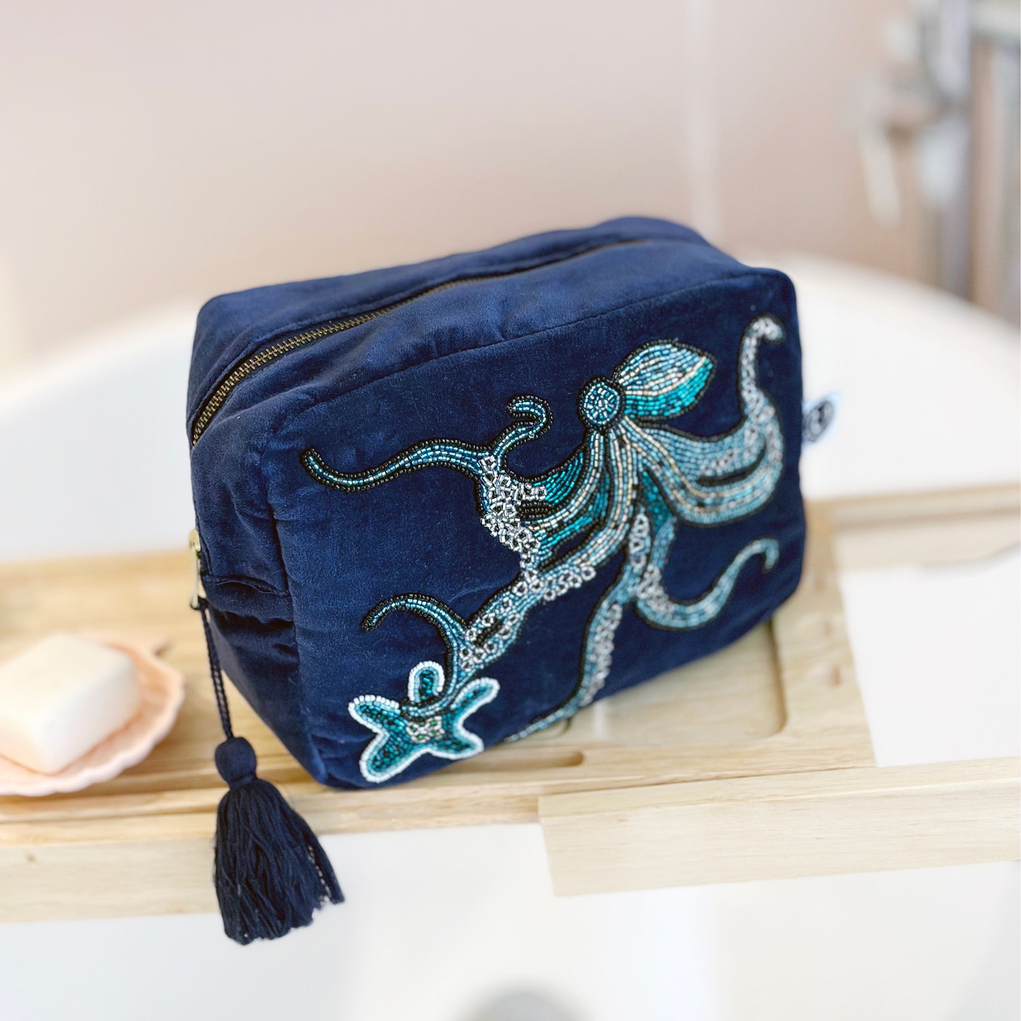 Beaded navy velvet wash bag with octopus and starfish on the front. The bag is on a wooden shelf on a bath and the taps are blurred in the background.