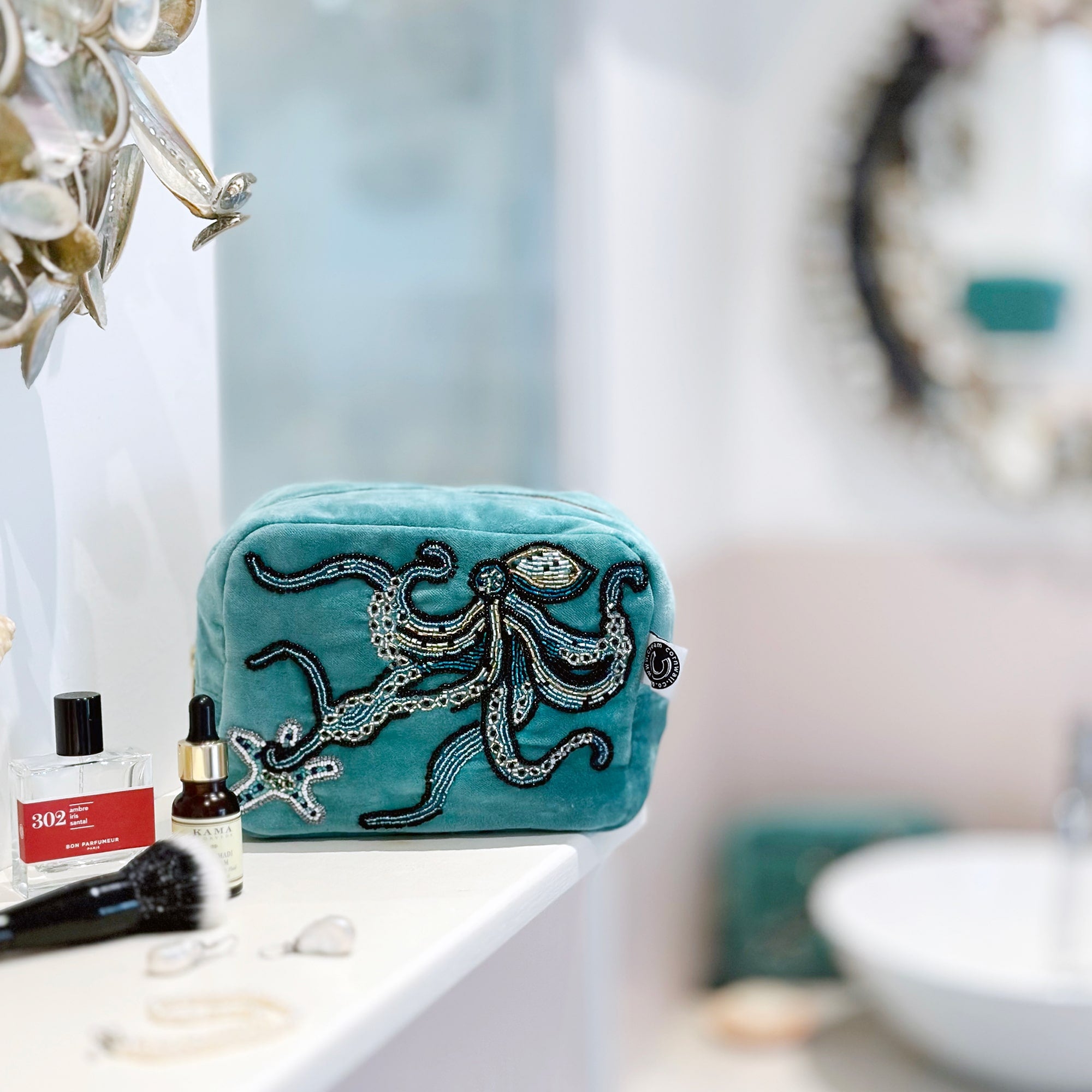 Beaded aqua velvet cosmetic bag with octopus and starfish decoration sitting on a bathroom shelf with a basin and shell mirror out of focus in the background.