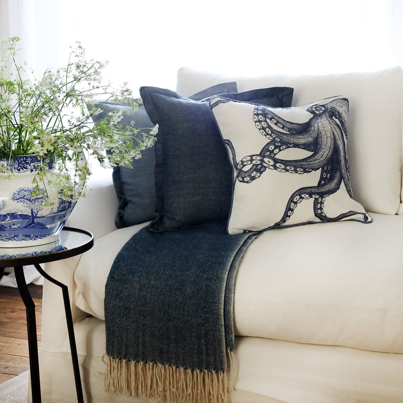 row of two blue cushions and a blue & white octopus cushion at the front, sitting on a navy throw on a white sofa with bright sunlight window behind and a large willow pattern jug filled with cow parsley
