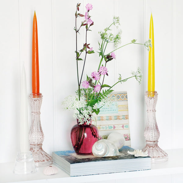 orange, yellow & white toned taper dinner candles in glass candle sticks, sitting on a white shelf with books and a cranberry glass jug with wild flowers