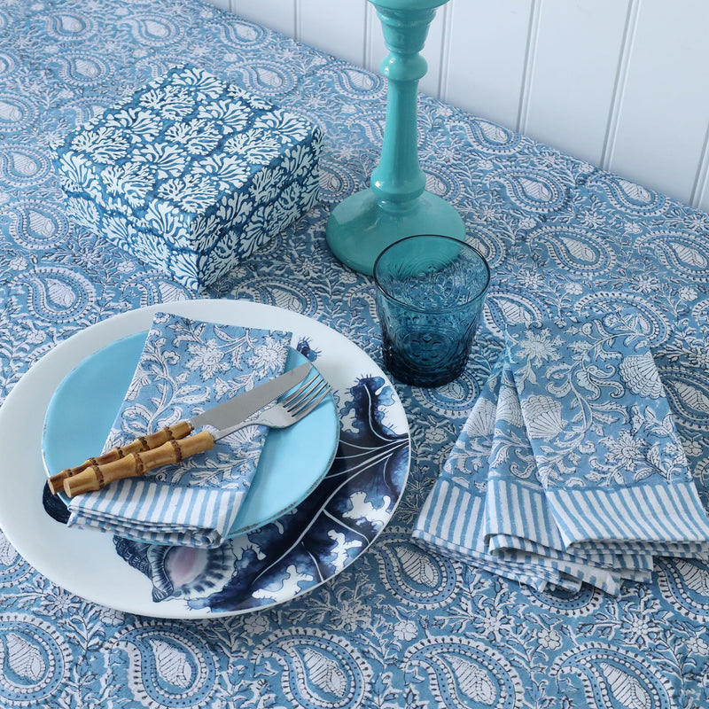 Azure Paisley Shell tablecloth which is a Hand block printed fabric in a soft blue paisley design.Placed on the tablecloth are beachcomber dinner plate,side plates and matching napkins.In the middle of the table is a blue glass,hand decorated box and a candlestick also in coordinating blues.