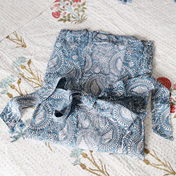 Azure Paisley Shell  dressing gown which is Hand block printed fabric in a soft blue and the print is swirling shells and tendrils in white.Placed on a bed with a white cover with occasional bright flowers.