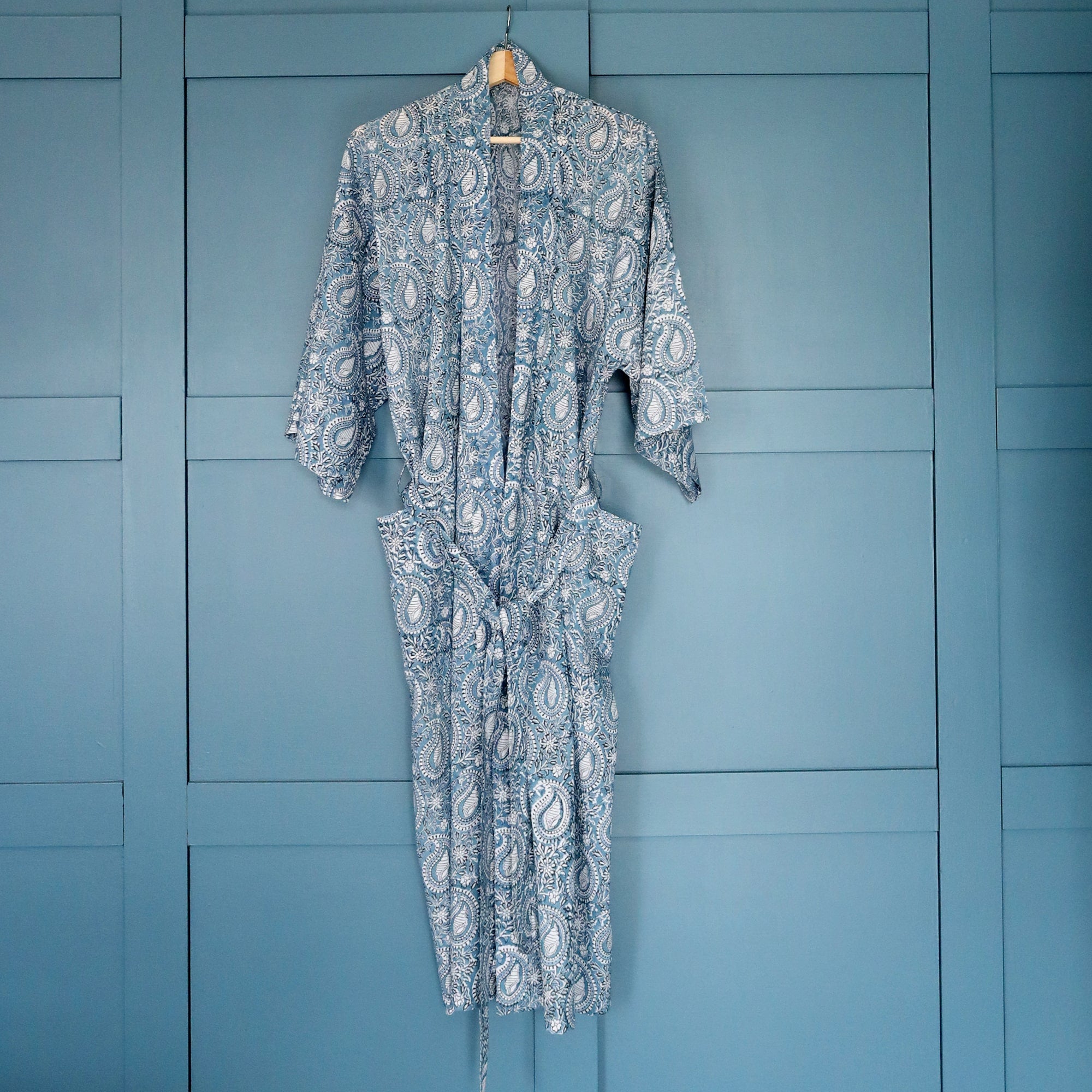 Azure Paisley dressing gown which is Hand block printed fabric in a soft blue  on a hanger in front of blue cupboards.
