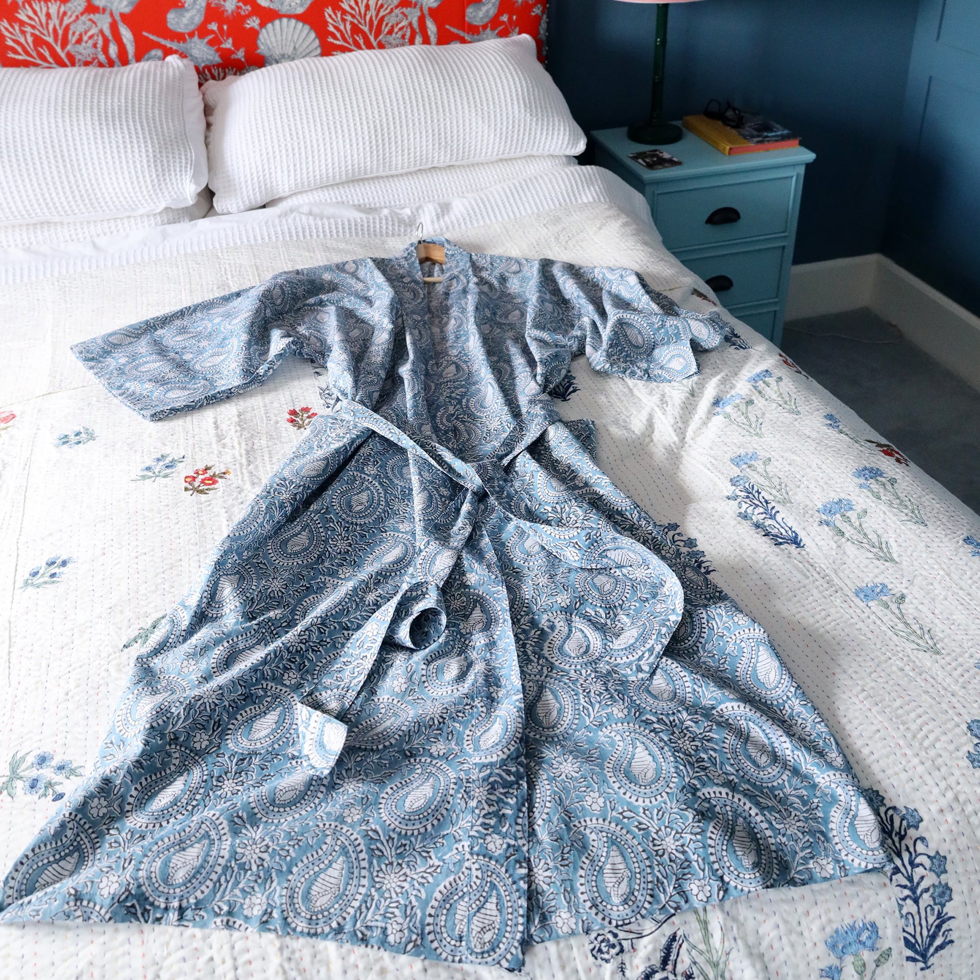 Azure Paisley Shell dressing gown which is Hand block printed fabric in a soft blue.At the head of the bed is a bright red and white headboard with shell patterns.The gown is placed on a bed with a white cover with occasional bright flowers. You can see the blue bedside cabinet with a lamp base and a marble swirl lampshade.