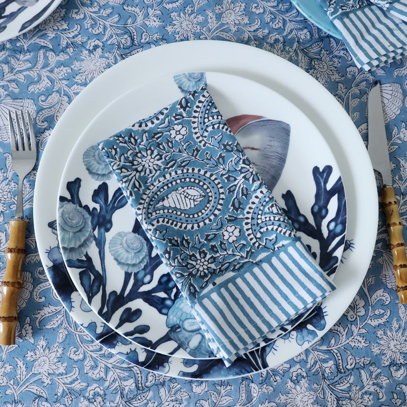 An Azure Paisley Shell napkin on Hand block printed fabric placed on beachcomber plate and side plate.On top is a set of bamboo cutlery and all placed on a matching Hand blocked tablecloth