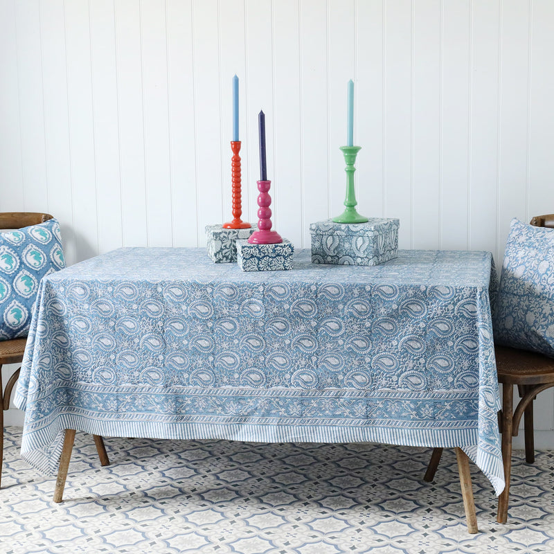 Azure Paisley Shell tablecloth which is a Hand block printed fabric in a soft blue and the print is swirling shells and tendrils in white .Around the pattern is a white and blue striped finished edge.It is placed on a rectangle table decorated with Hand decorated boxes and candlesticks.Either side of the table are chairs with a hand block printed cushion.