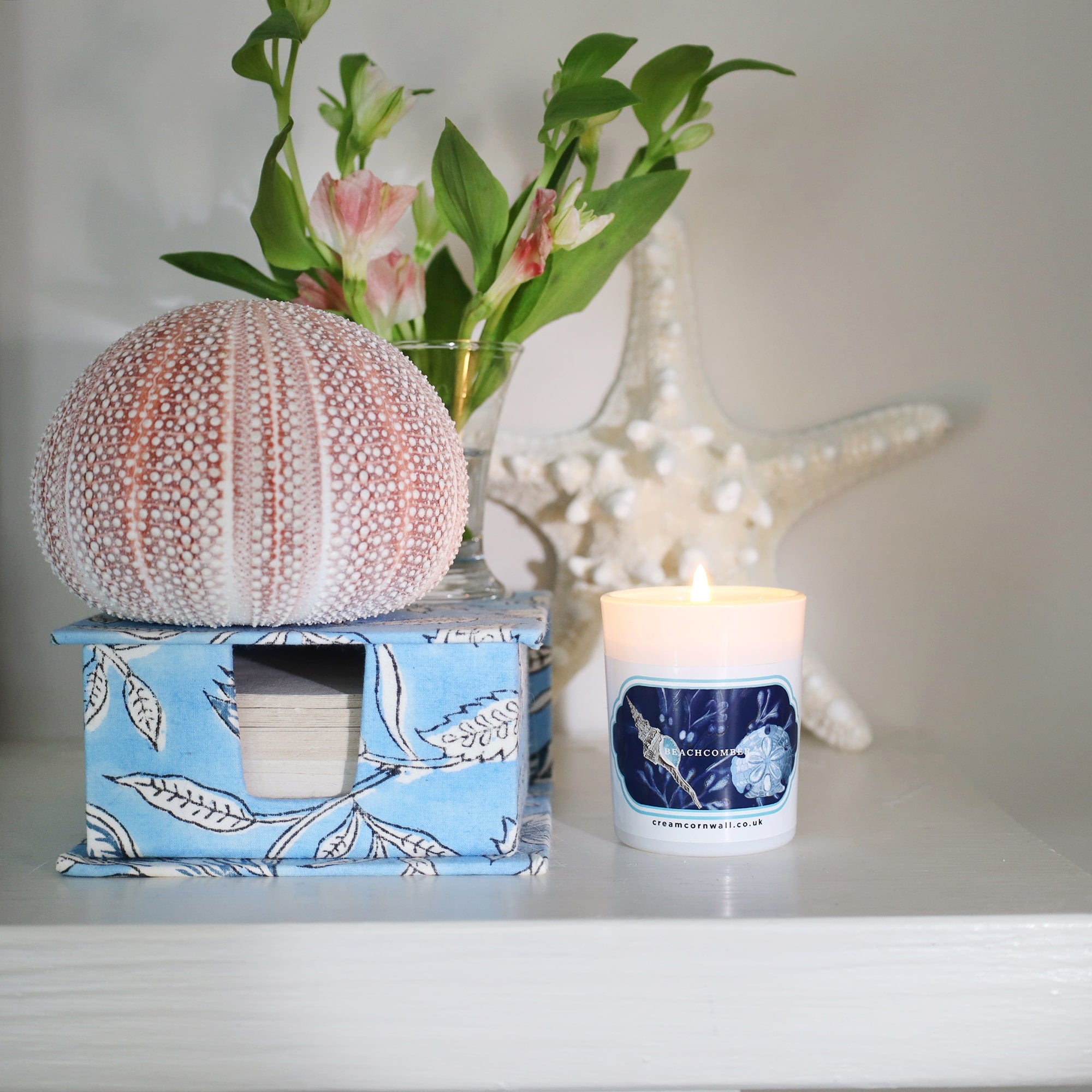 small candle with shell design sticker on the front with flowers, sea urchin and starfish on a shelf