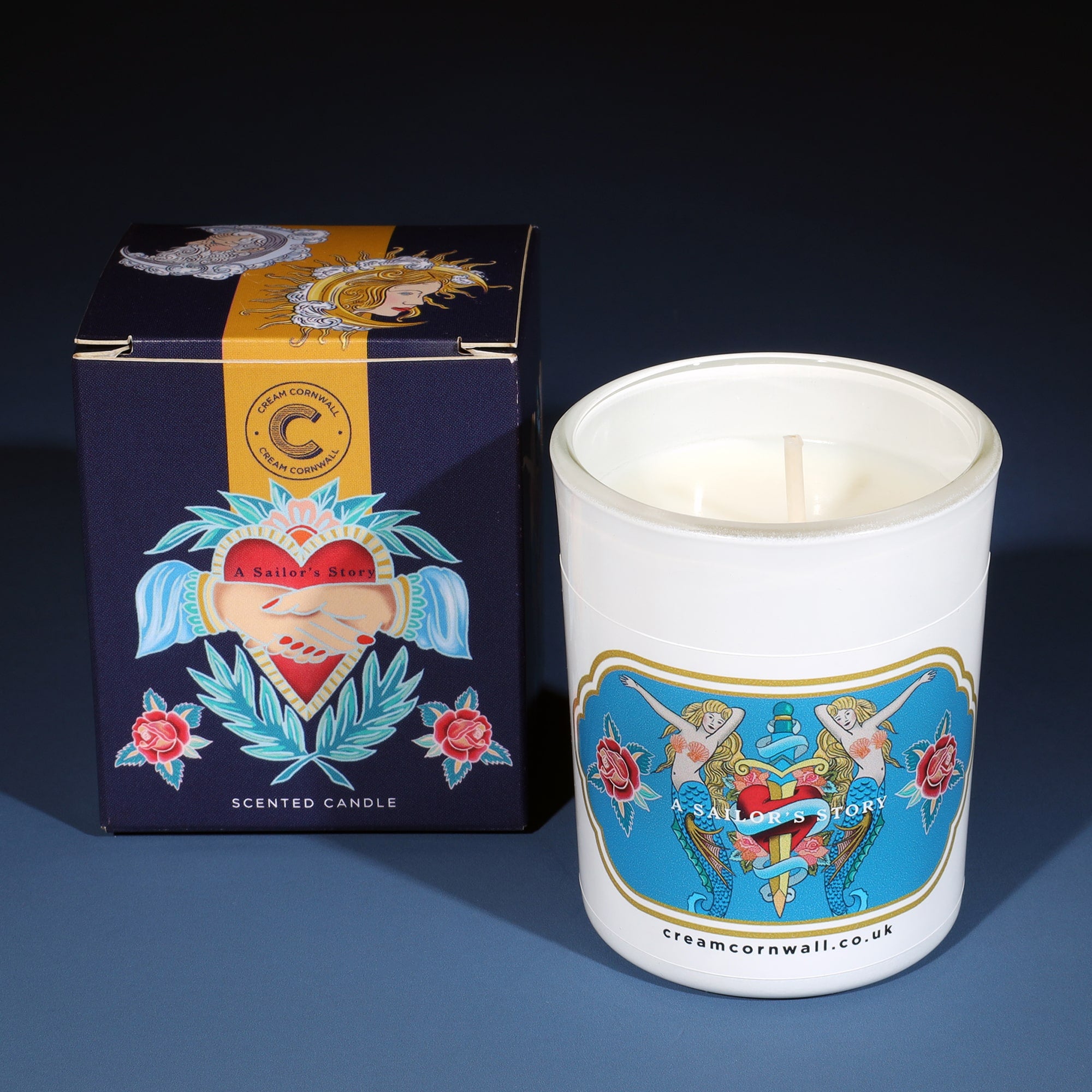 Small candle with box on graduated navy blue background. the box and candle have tattoo designs based on sailor's tattoo's.