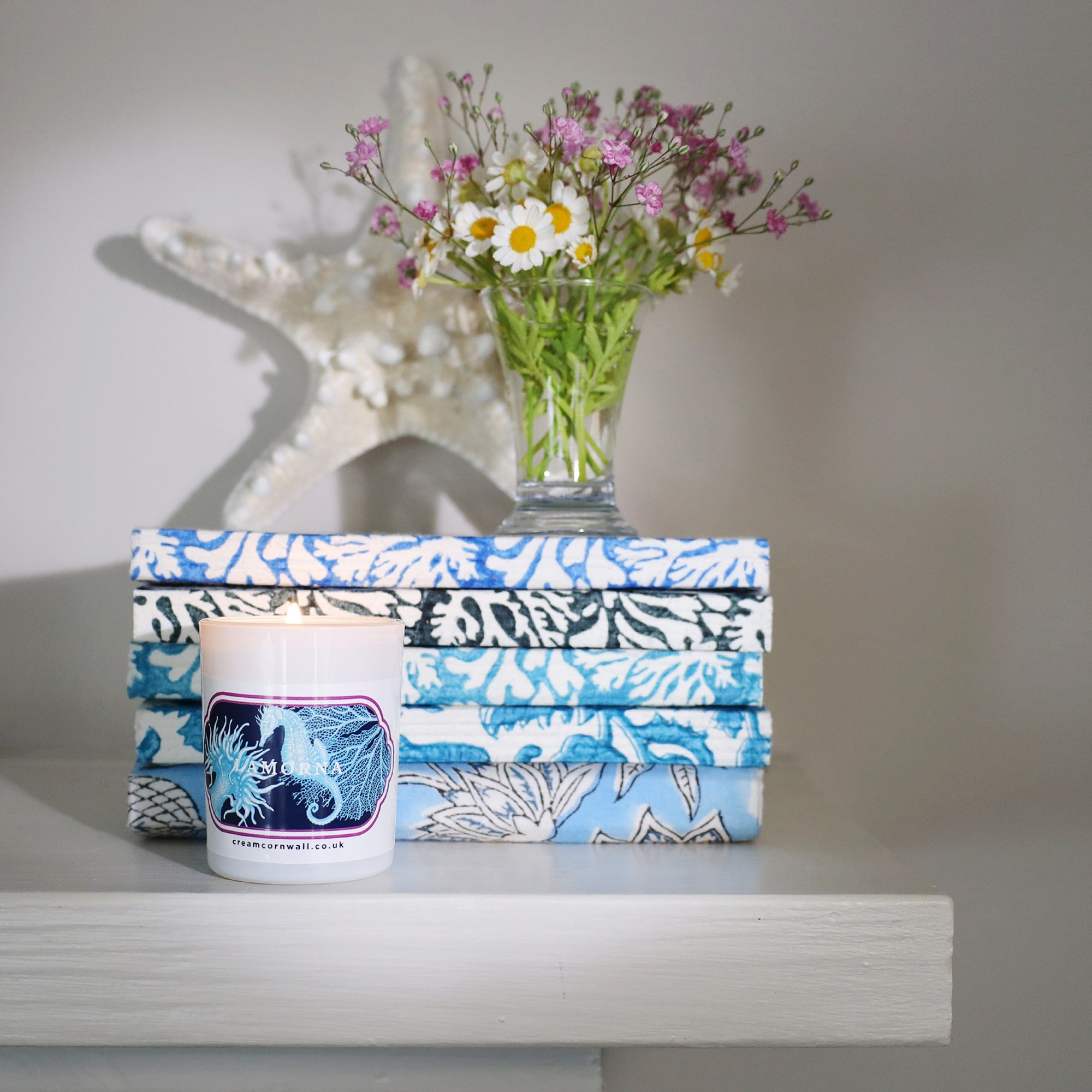 Small candle with seahorse and coral design sticker on the front, on a shelf in front of pile of blue notebooks with a small vase of flowers and a starfish behind.