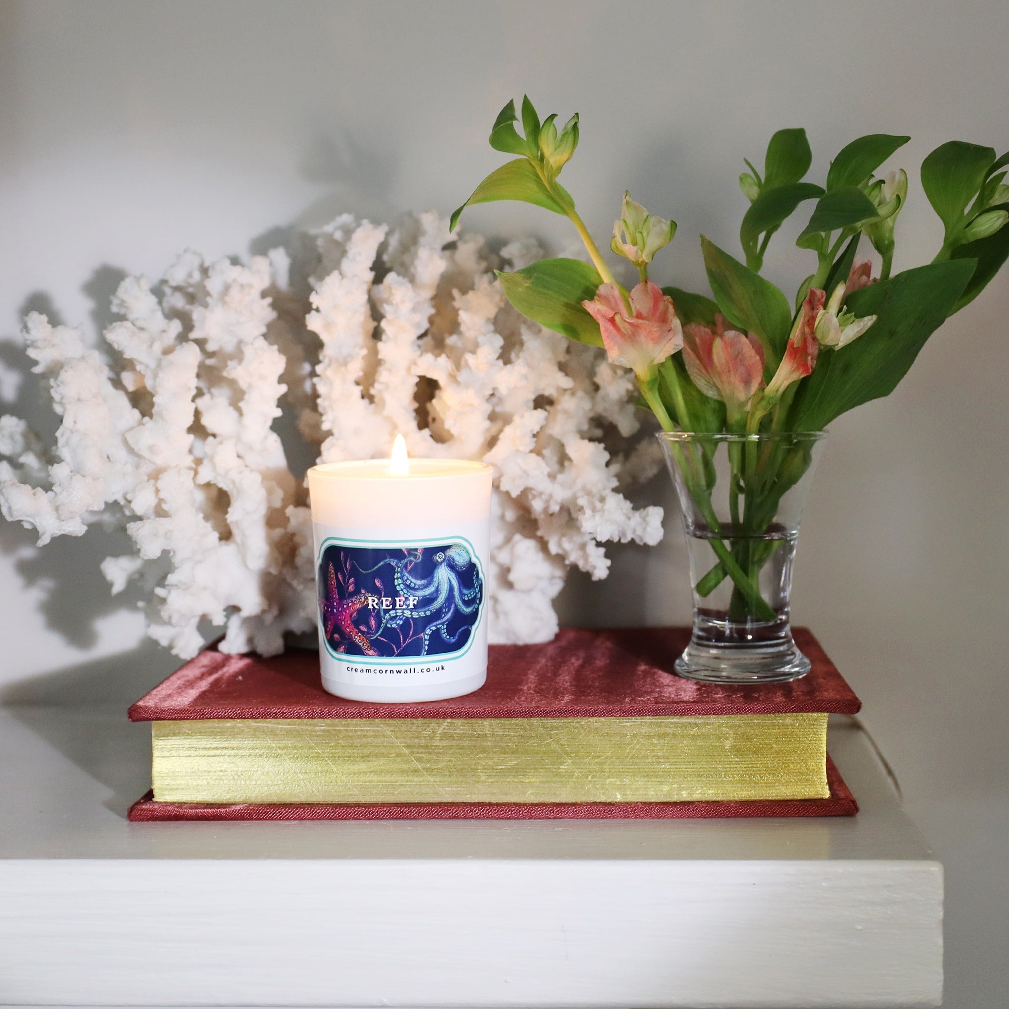 Small Reef candle in white votive with colourful sticker of sea creatures on the front. This is sitting on a book with some coral behind a small vase of flowers to the side.