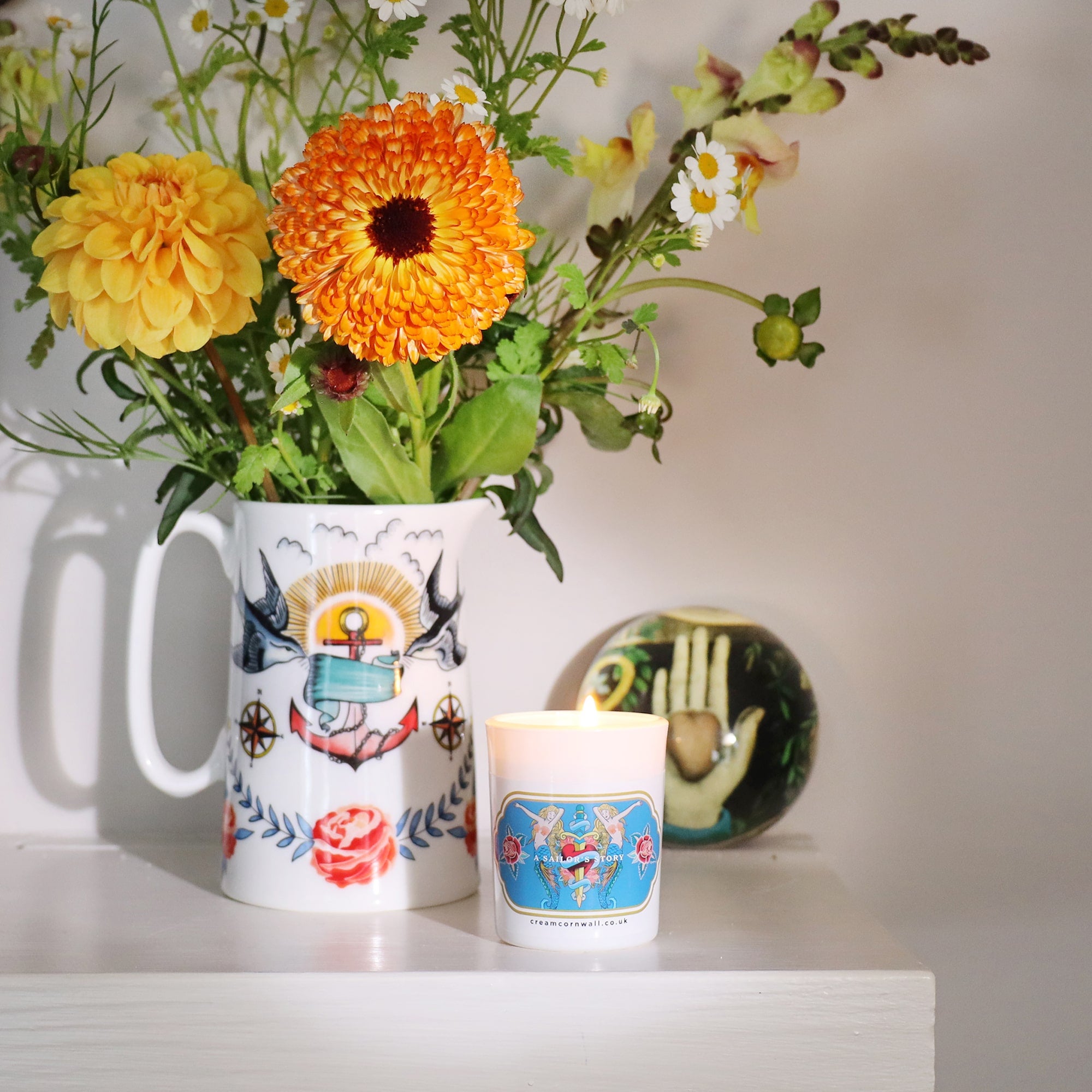 Small travel candle on shelf with mermaid tattoo design on the front. This is sitting on a shelf with a jug of flowers with complimentary design and a paperweight in the back ground.