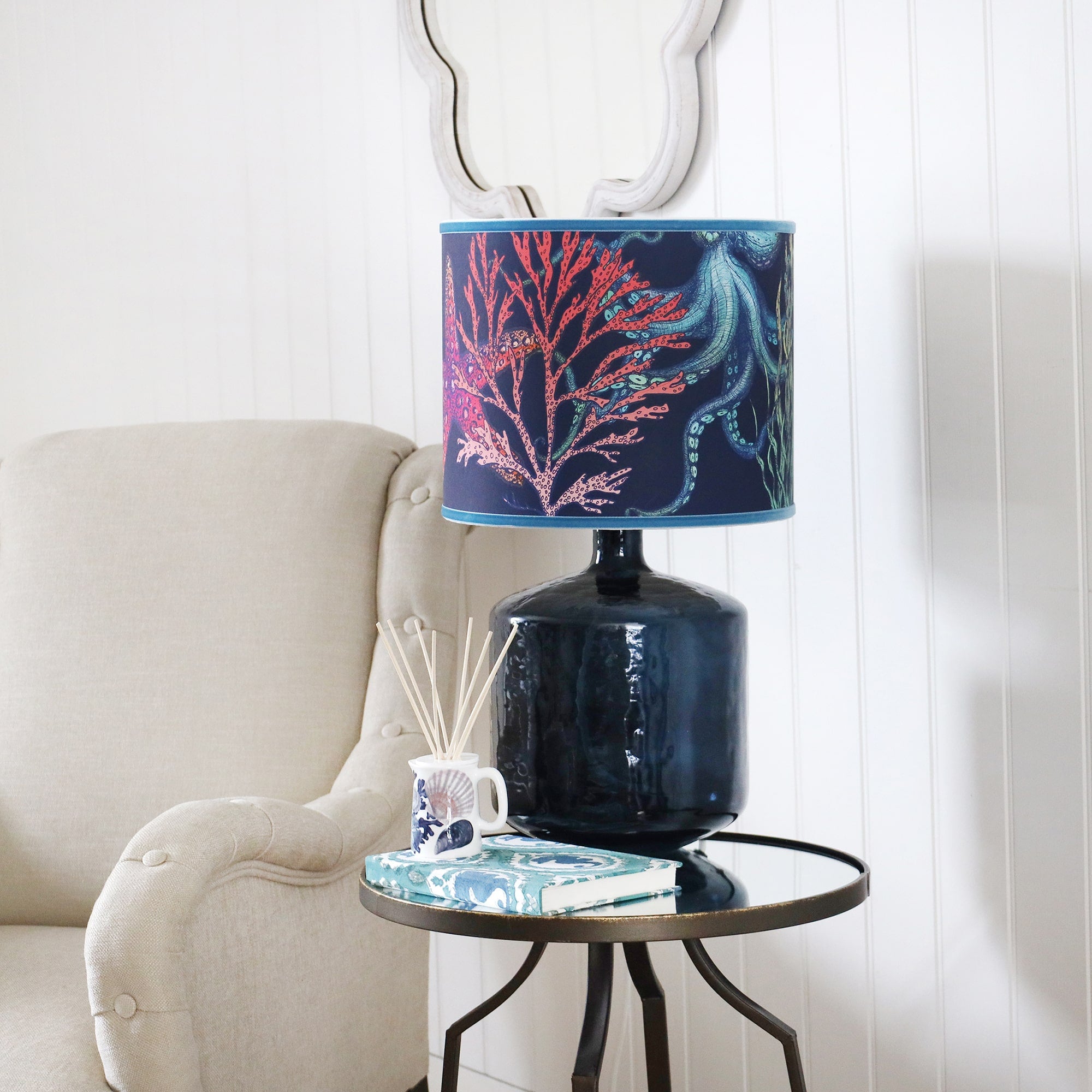 Zennor Glass lampbase with a Navy Reef lampshade on a mirror table.On the table is a  Beachcomber diffuser and a notebook.Next to the table is a chair on the wall behind is a wave mirror.