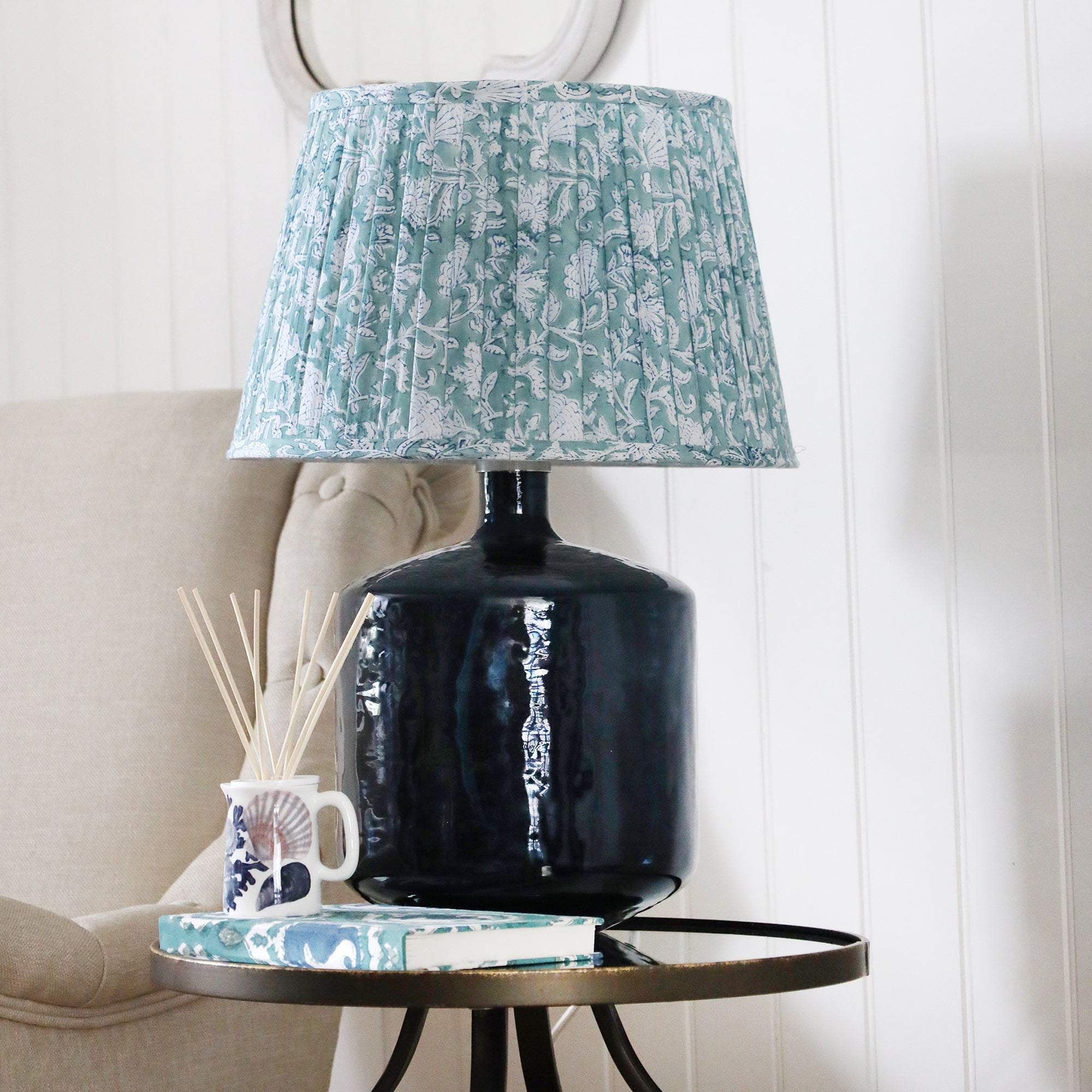 Zennor Glass lampbase with a pleated lampshade on a mirror table.On the table is a Beachcomber diffuser with a notebook.Next to the table is a chair, on the wall behind is a wave mirror.