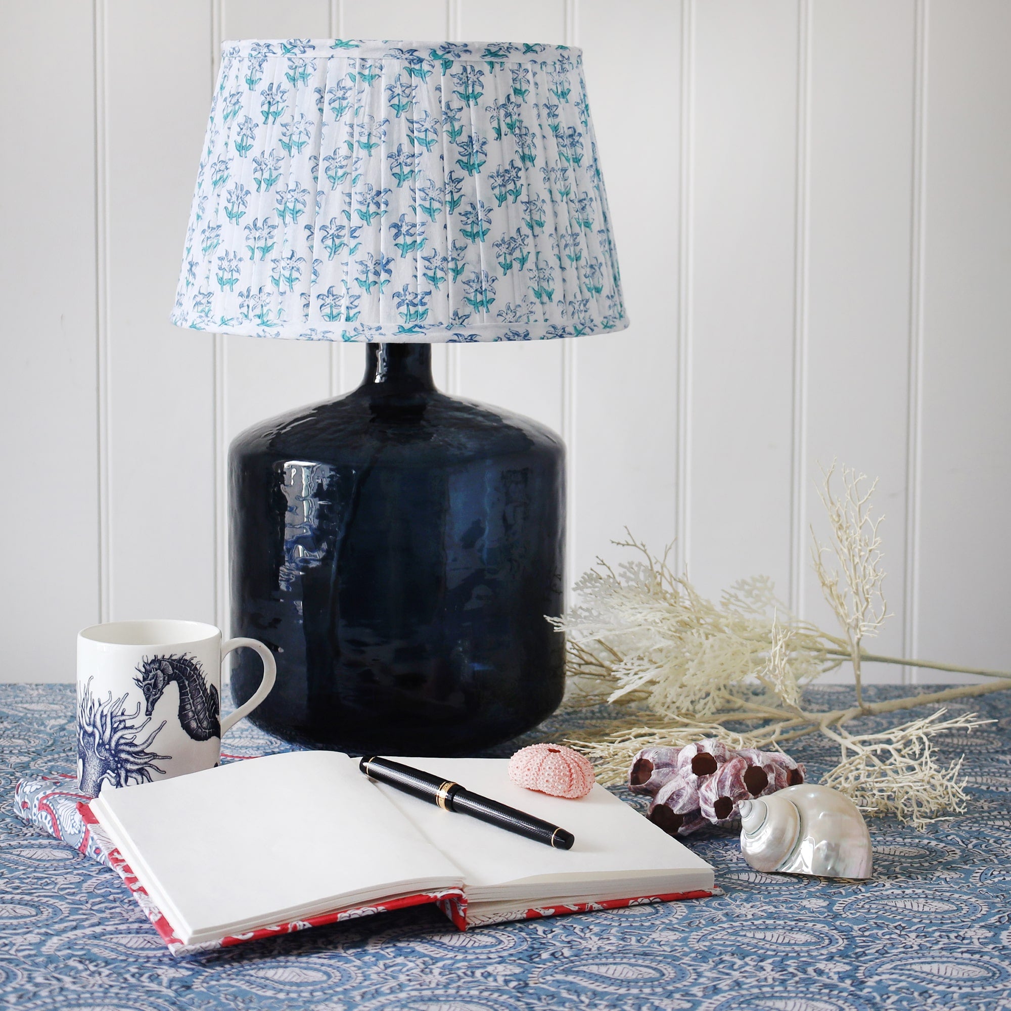 Medium pleated lampshade in hand blocked print in blue and white in our seastar design on a dark blue glass lampbase placed on a table.On the table is notebooks and a pen next to a seahorse mug and some fauna next to the lampbase