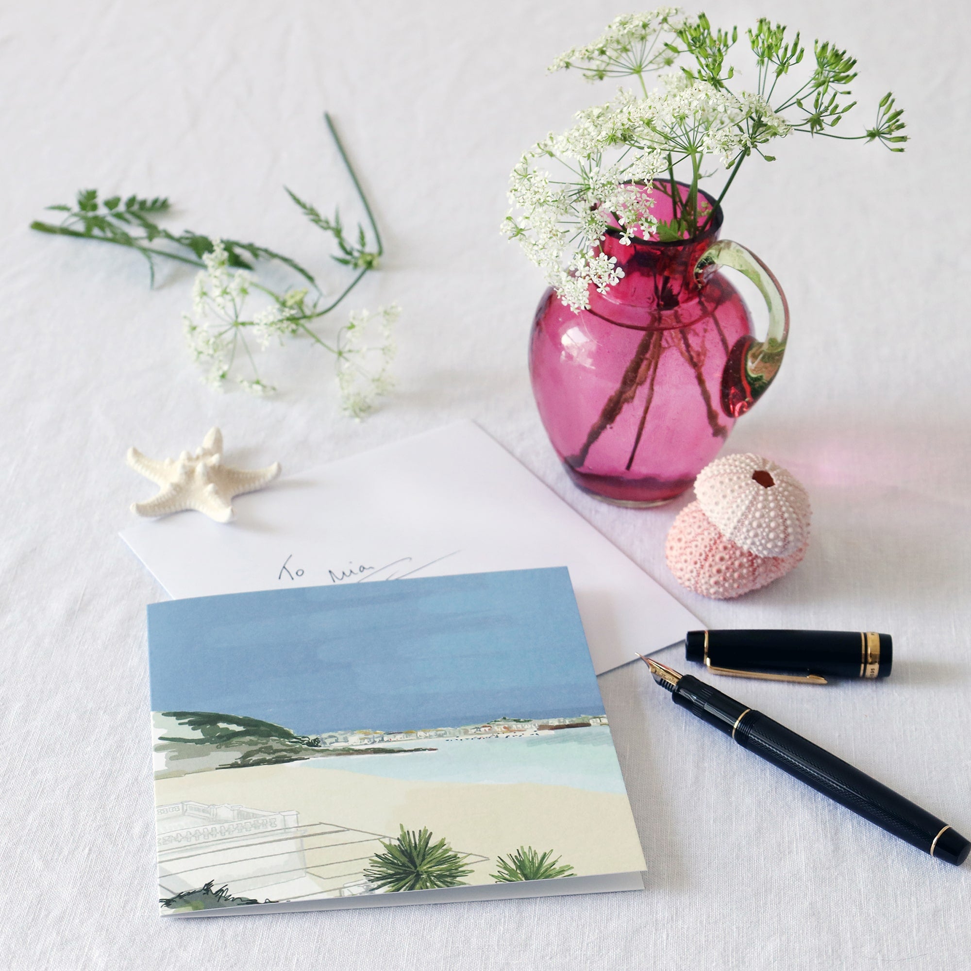 greeting card with illustration of the view across the beach from porthminster cafe st ives lying on a white table cloth with a fountain pen, hand written envelope shells and a small cranberry glass jug with wild flowers in 