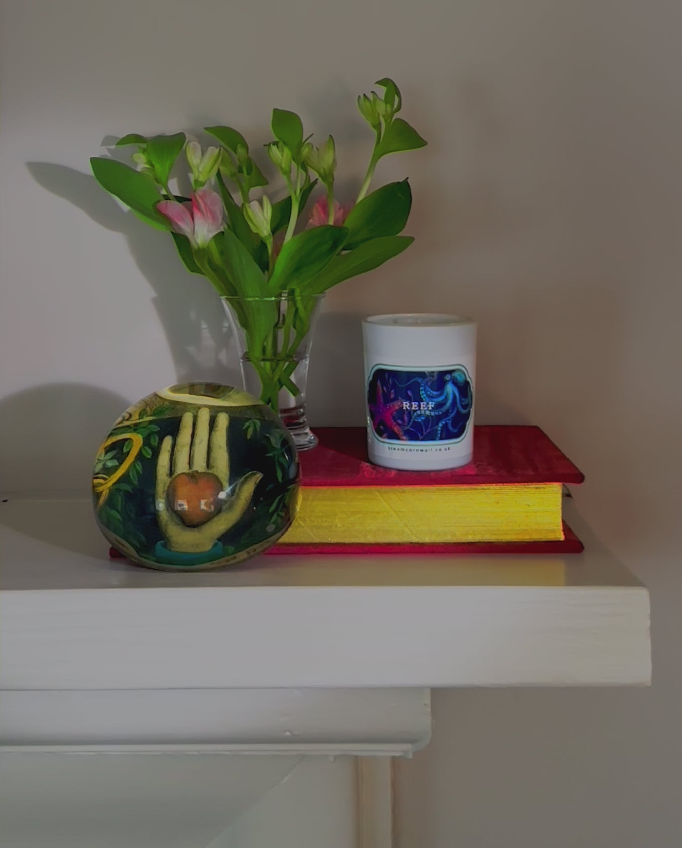 Small candle with brightly coloured sticker of sea creatures on the front, sitting a book with w small vase of flowers and paperweight & the candle is being lit with a match.