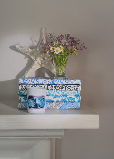 Small candle with seahorse and coral design in front of stack of blue notebooks, a small vase of flowers and a starfish. The candle is being lit  with a match