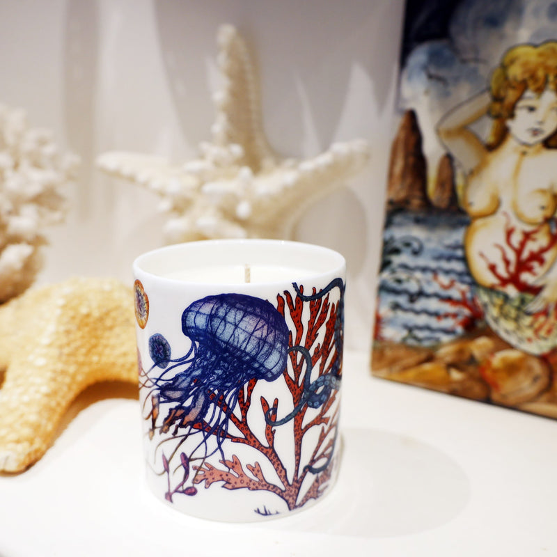 Candle in a bone china beaker decorated in brightly coloured sea creatures, coral and seaweed on a shelf with starfish on the background and part of a hand painted tile with a mermaid on it just in focus.