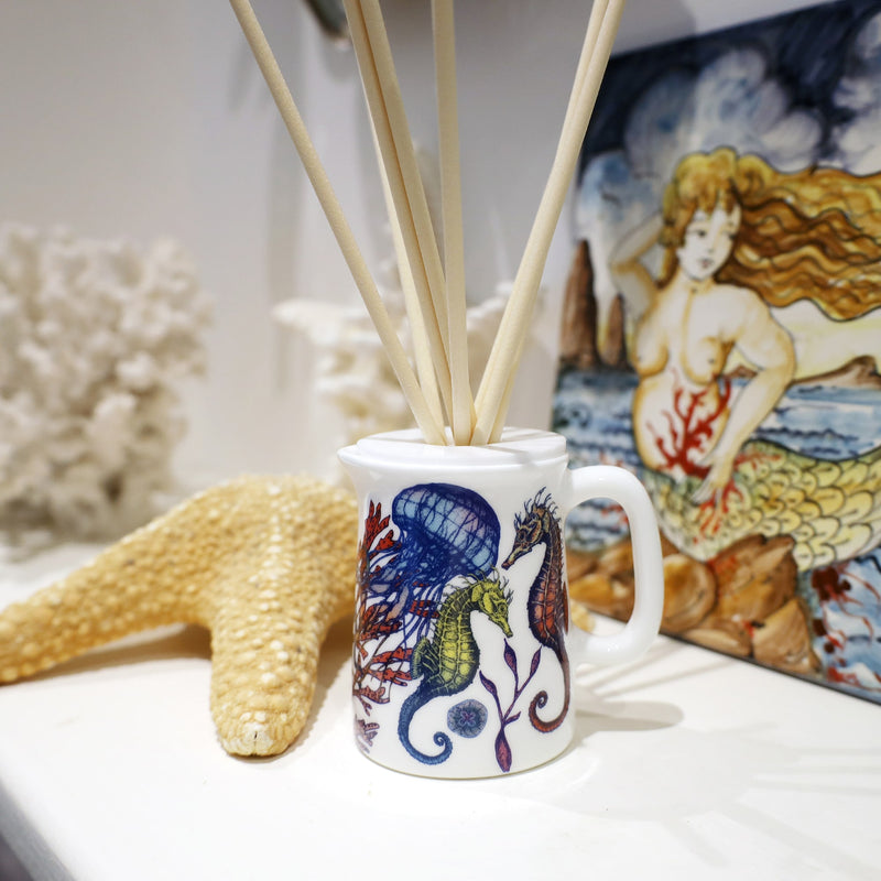 A small bone china jug used as a room diffuser with reeds coming out of the lid. It is decorated in brightly coloured sea creatures, coral and seaweeds. This is sitting on a white shelf with corals and starfish and a hand painted tile of a mermaid sitting on a rock.