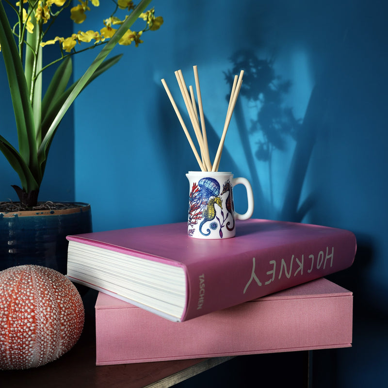 Bone china room diffuser in a small jug with colourful octopus, coral and seaweed design sitting on a 2 pink books against and tea blue wall with the sunlight reflecting a potted orchid on the wall behind.